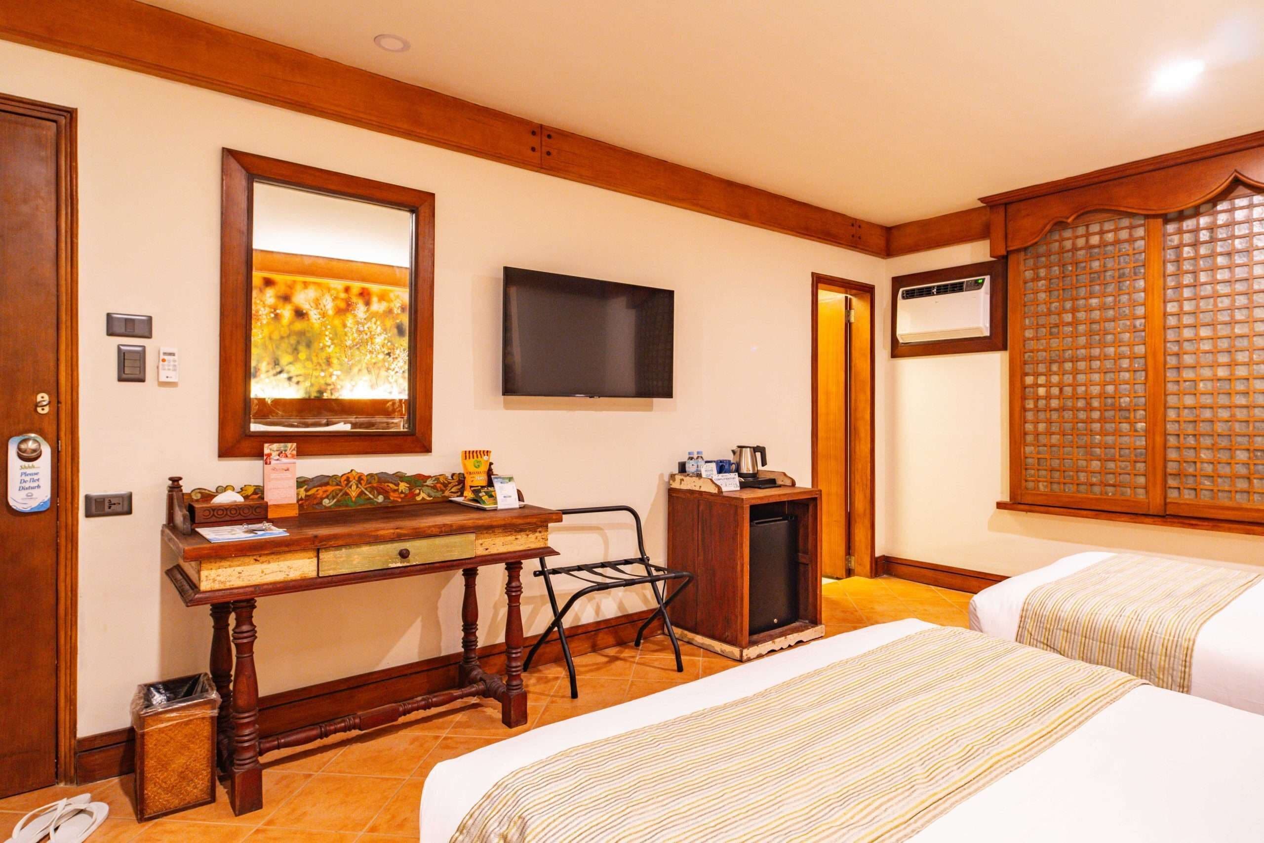 A hotel room with a wooden desk, a television, a minibar, a bed, and a traditional Asian-style window.