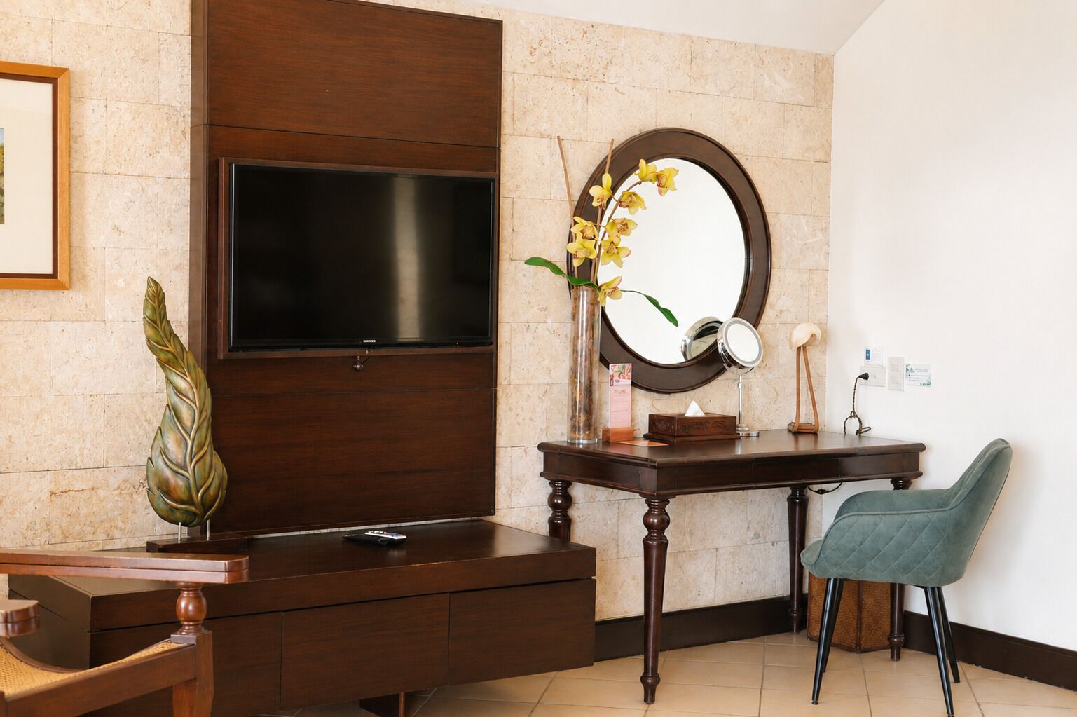 Une salle de bain moderne avec un meuble-lavabo en bois, un téléviseur à écran plat fixé au-dessus, un miroir rond et un vase avec des fleurs.