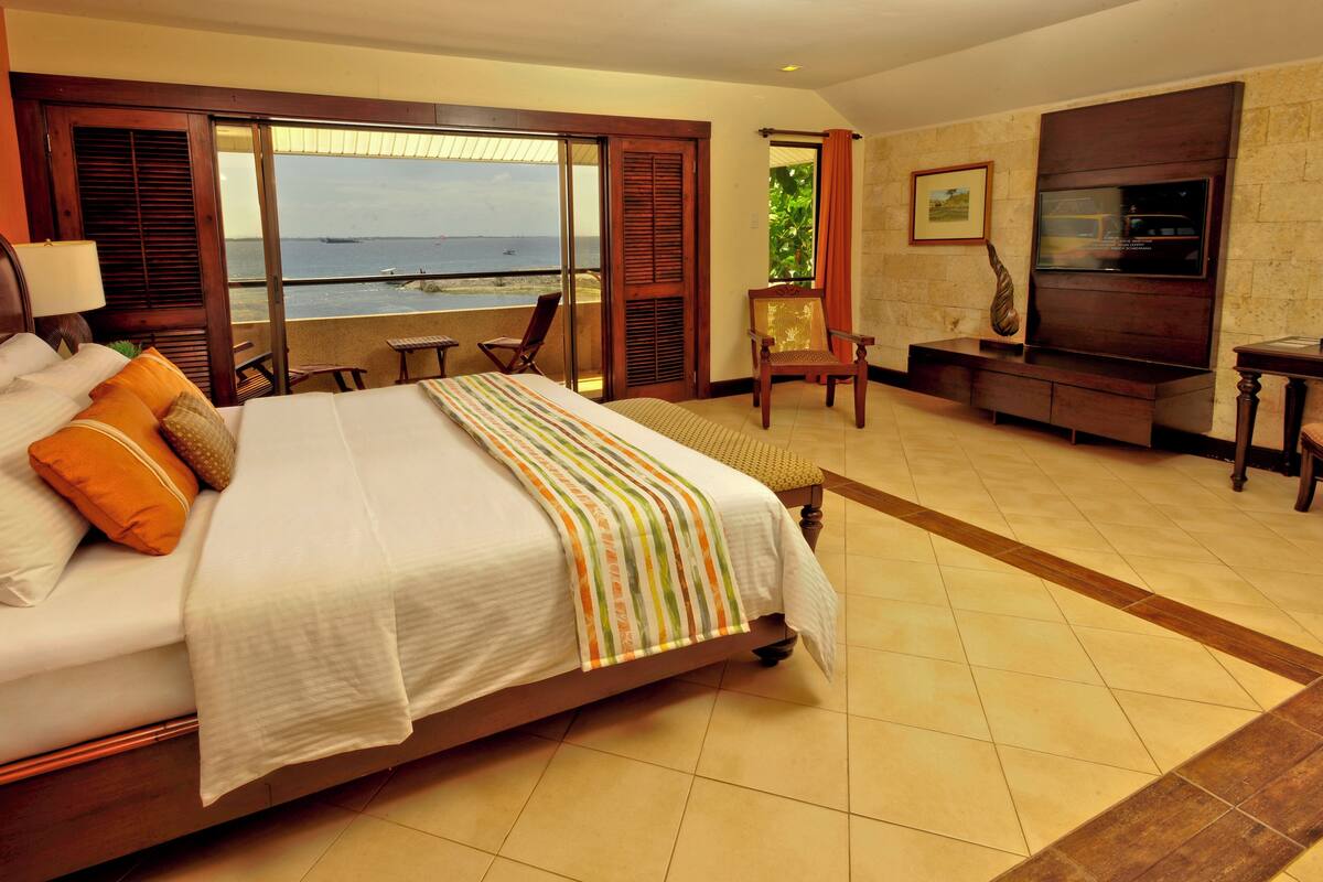 Quarto de hotel com uma cama grande, televisão, poltrona, mesa pequena e vista para o mar.