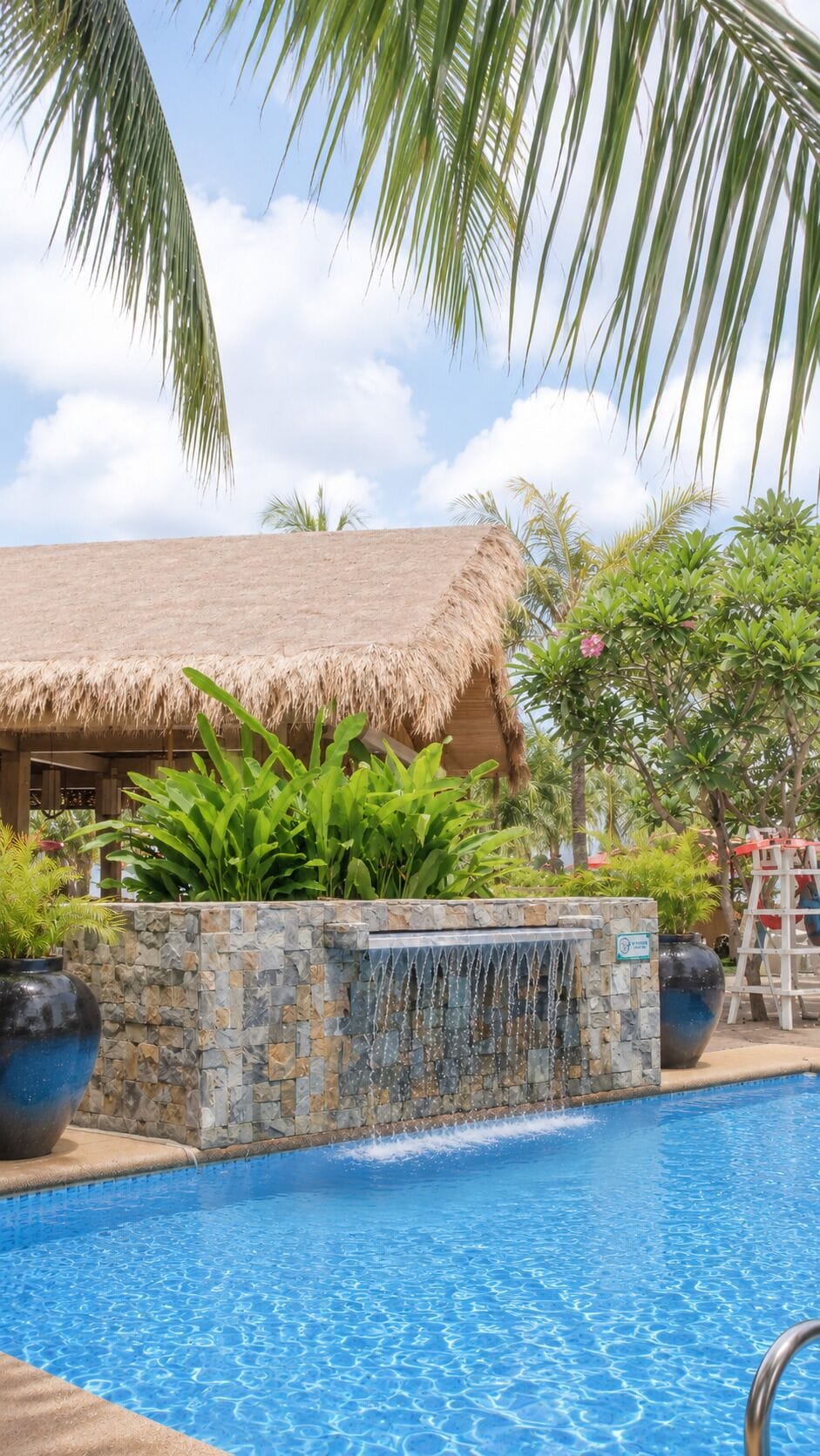 A swimming pool with a stone wall and a waterfall feature, surrounded by tropical plants and a thatched-roof structure.