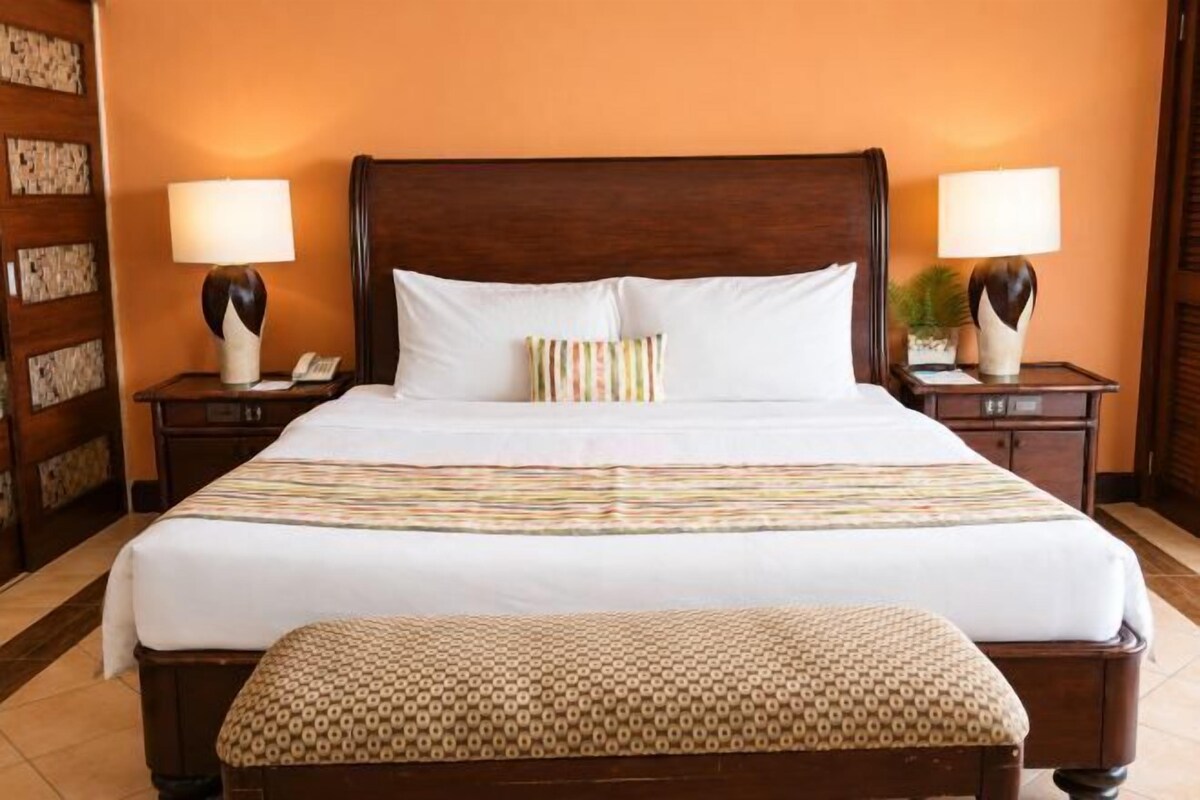 A hotel room with a large bed, two bedside tables with lamps, a wooden headboard, and a patterned bench.