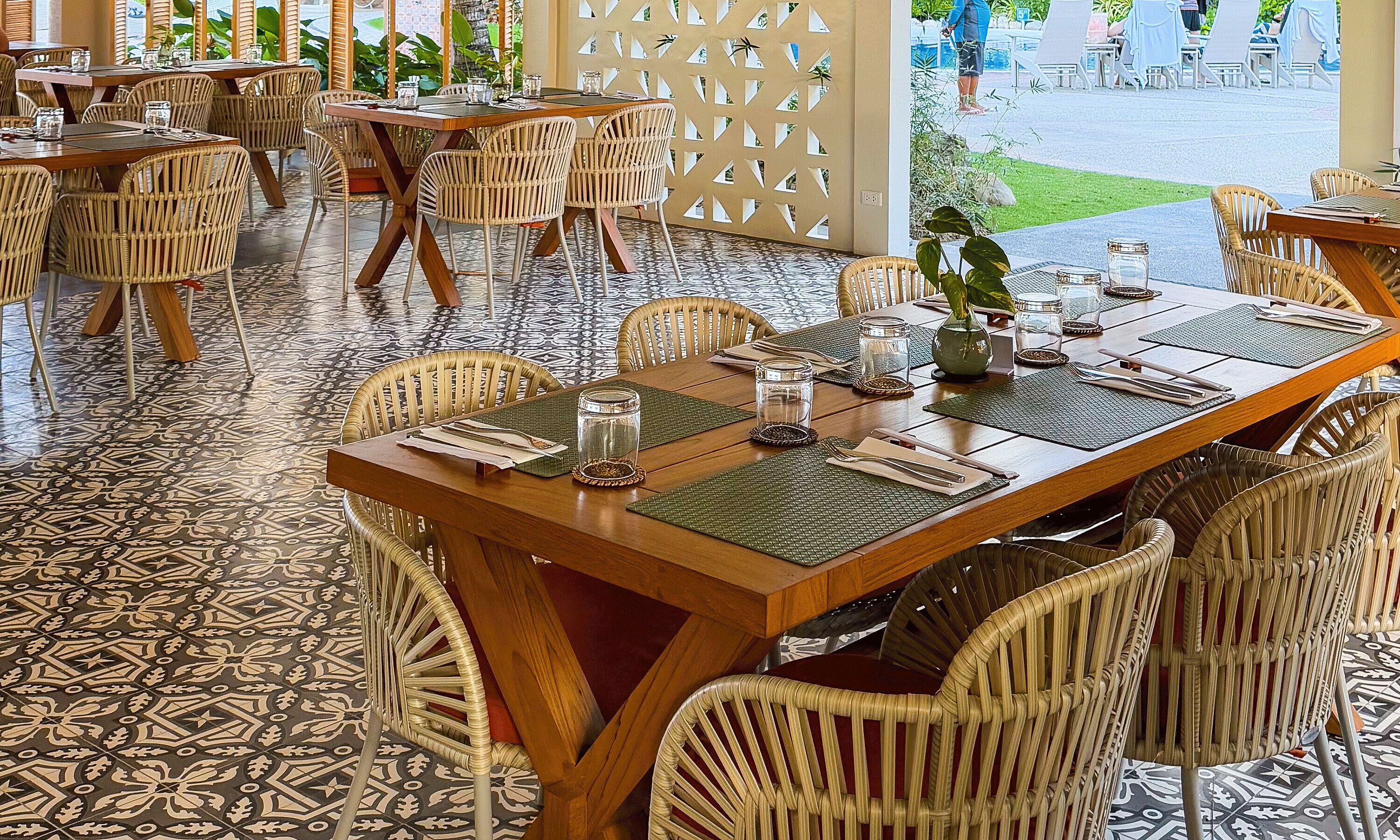 A restaurant with wicker chairs and tables, patterned floor tiles, and a view of a pool area.
