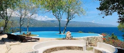 A pool area with a curved edge, surrounded by trees and a view of the sea.