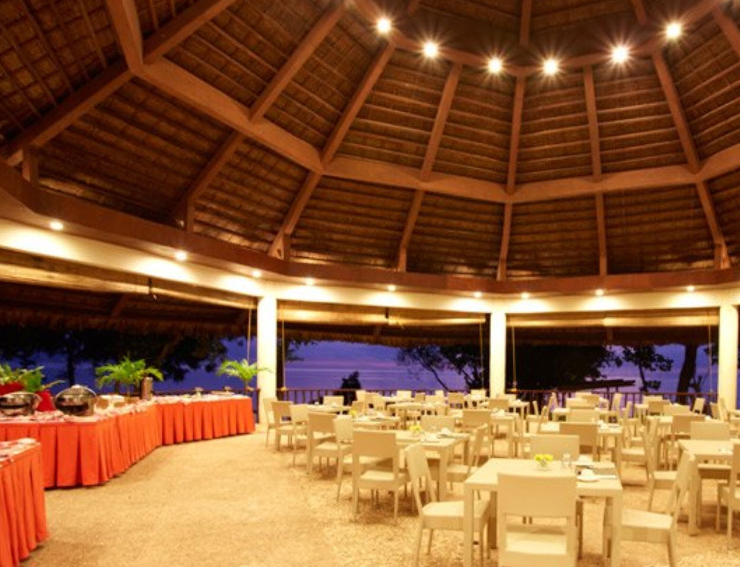 A large indoor dining area with a high ceiling and natural light. Tables are arranged in rows, and there are plants along the walls.