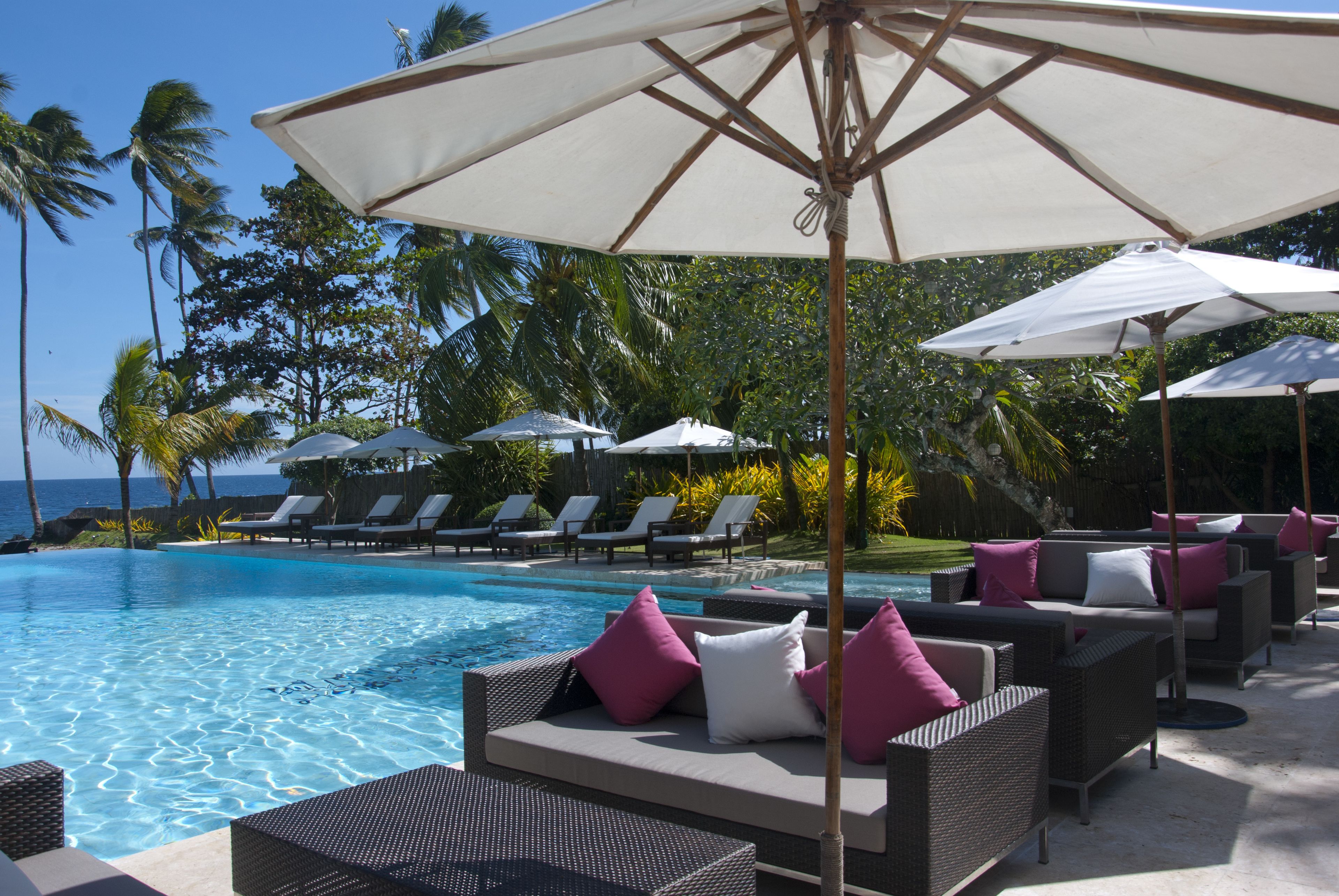 A poolside area with lounge chairs and umbrellas, surrounded by palm trees and a view of the ocean.