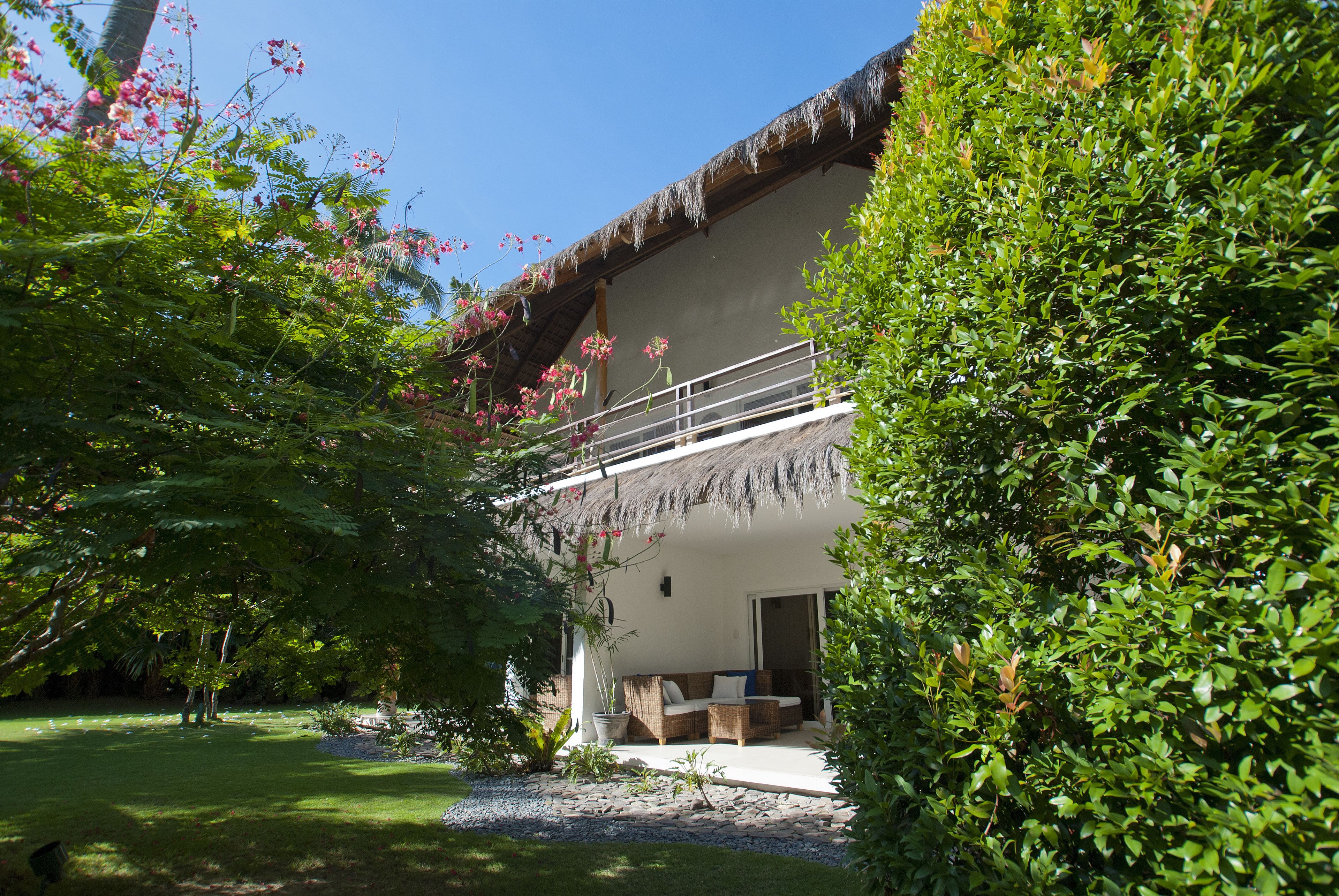 A two-story building with a thatched roof, a balcony with white railings, and a garden area with a lawn and potted plants.