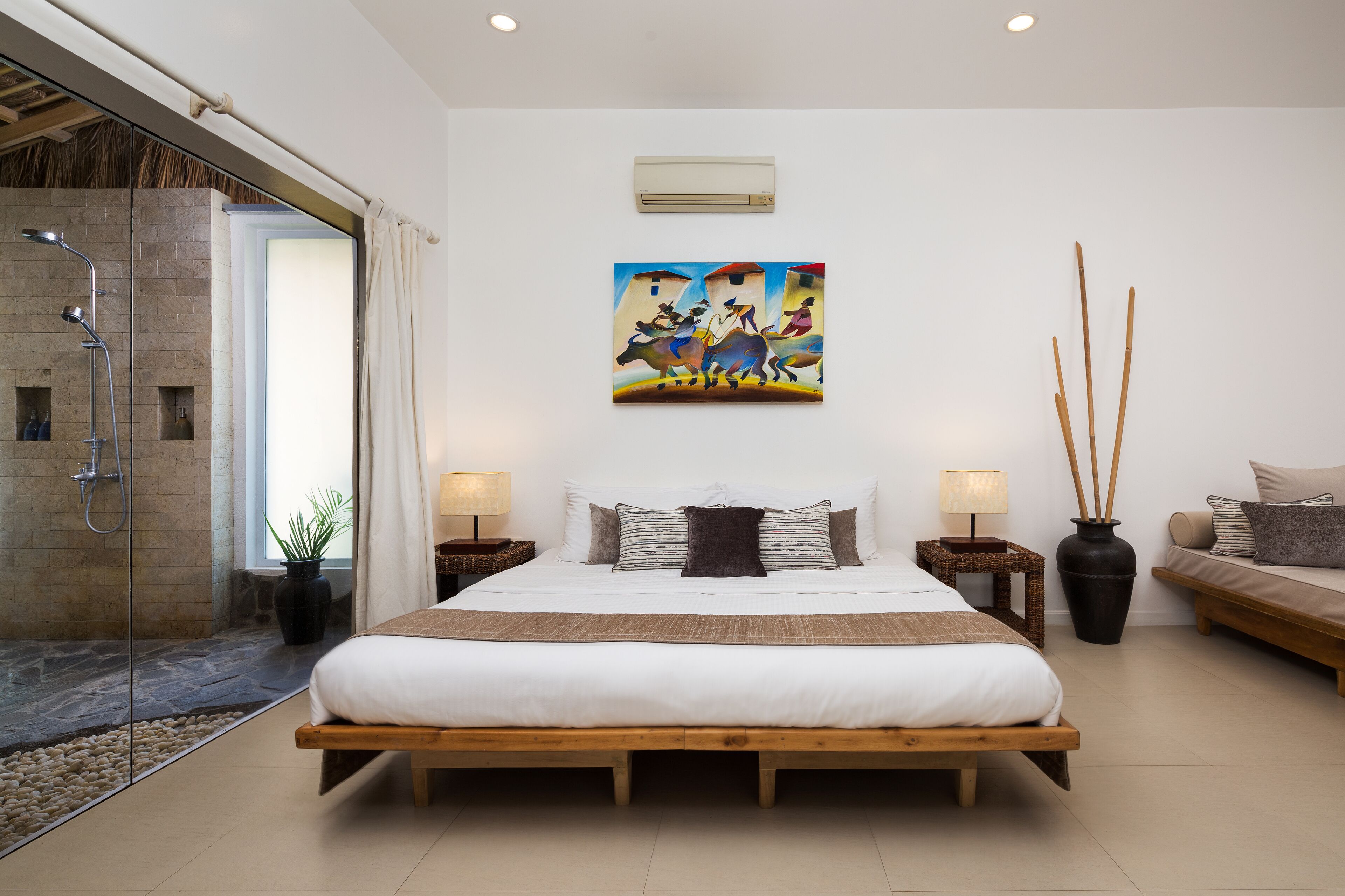A modern bedroom with a large bed, a shower area, a painting on the wall, and a seating area with a sofa and side tables.
