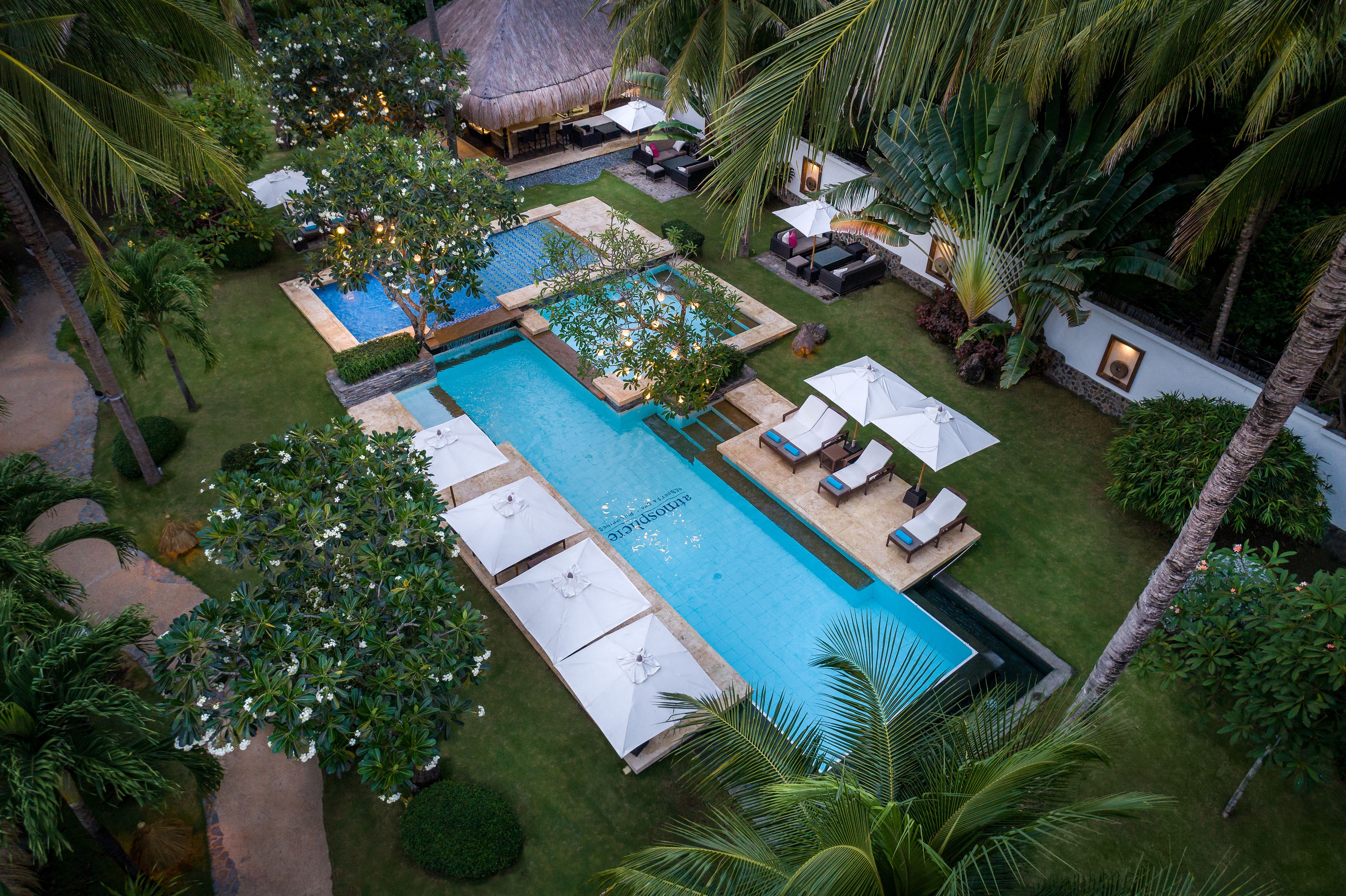 A large outdoor swimming pool with multiple lounge areas and umbrellas, surrounded by lush greenery and palm trees.