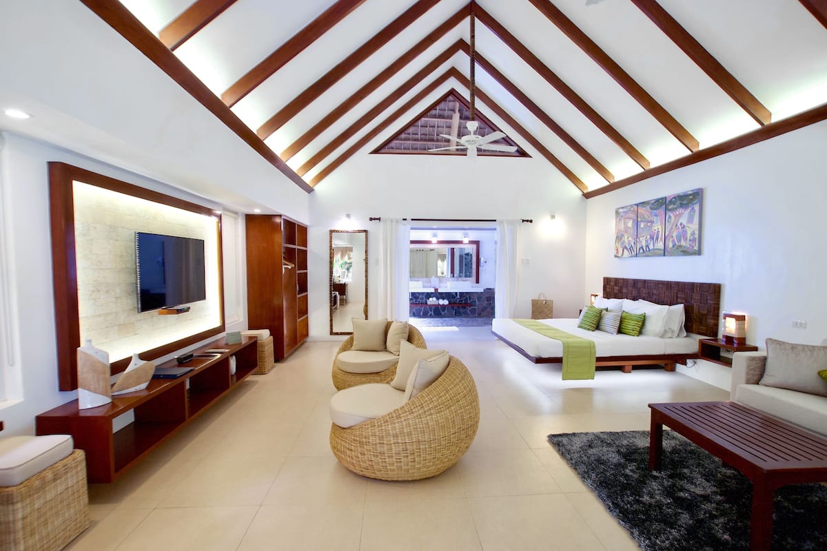 A spacious living area with a large flat-screen TV, a round wicker sofa, a wooden coffee table, and a bed with a green throw blanket.