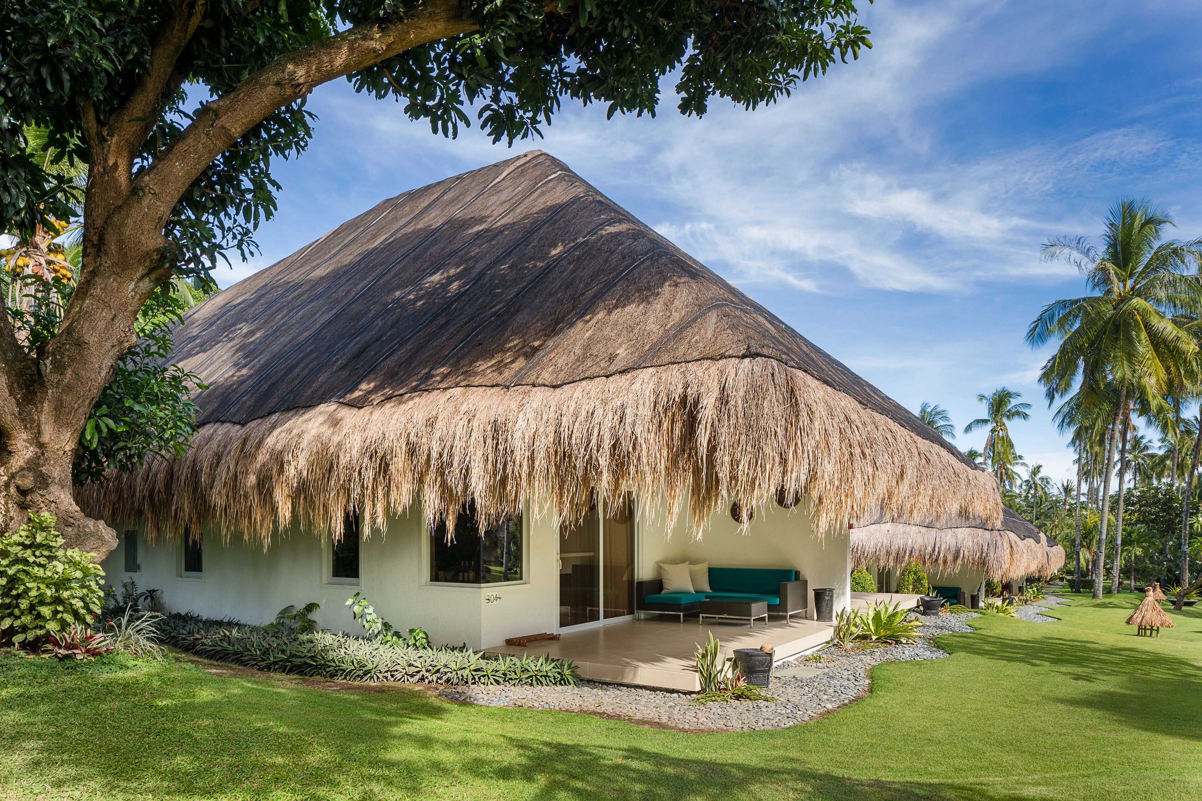 Een rieten huis met een overdekte veranda, omringd door weelderige begroeiing en palmbomen.
