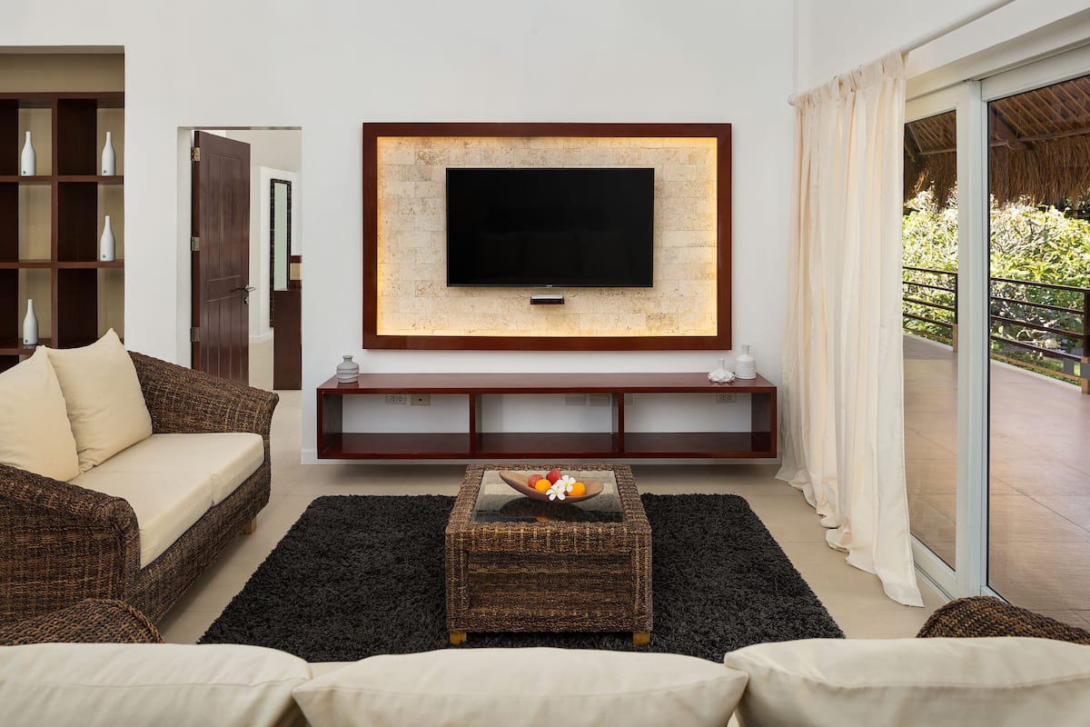 A living room with a flat-screen TV mounted on the wall, a wicker coffee table with fruit, a sofa, and a glass door leading to an outdoor area.