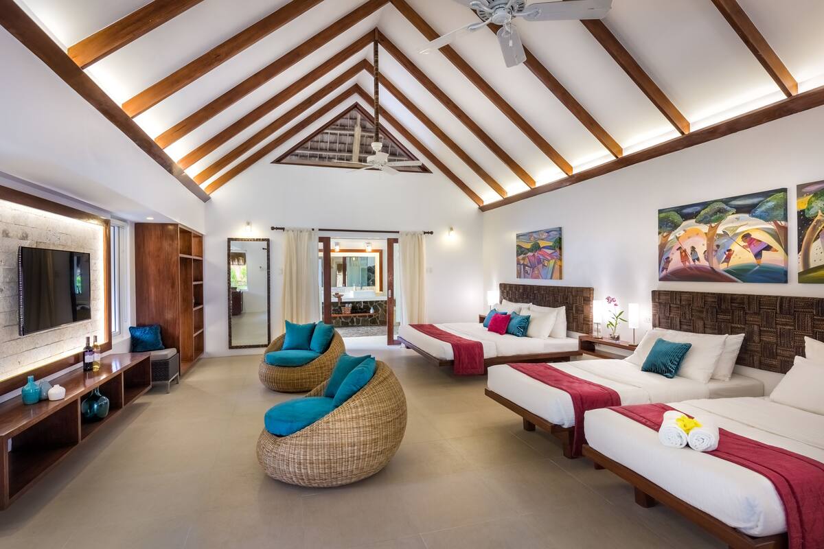 A spacious room with a high ceiling, wooden beams, two beds with red and white bedding, a large flat-screen TV, wicker chairs, and artwork on the walls.