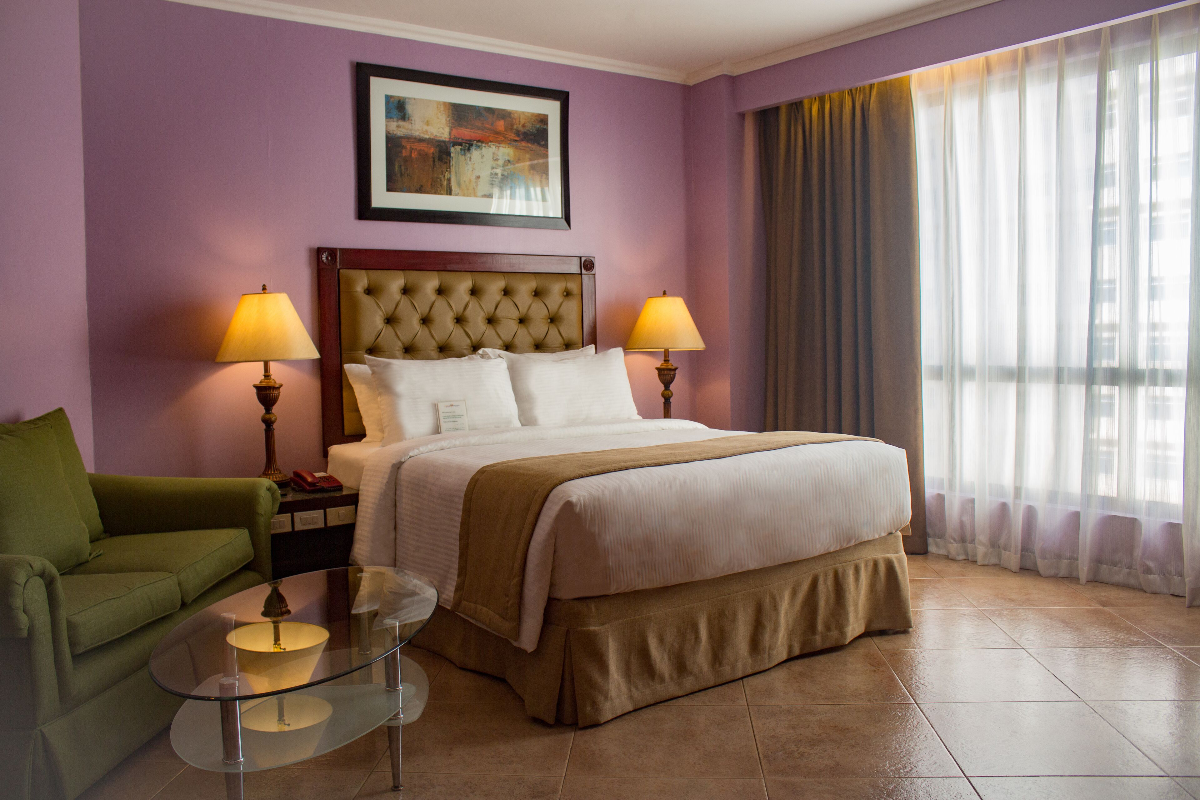 A hotel room with a bed, two bedside lamps, a green armchair, a glass coffee table, and a painting on the wall.