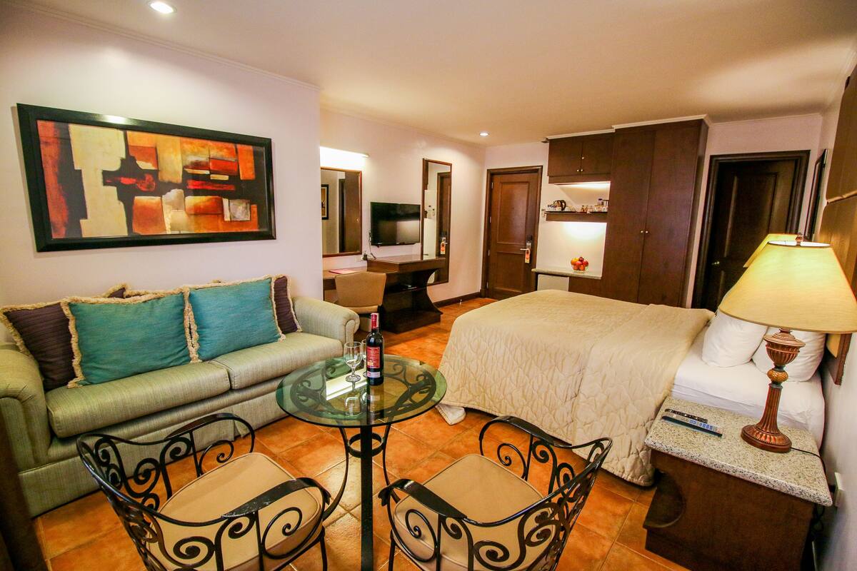 A hotel room with a bed, a sofa, a dining table, and a TV.