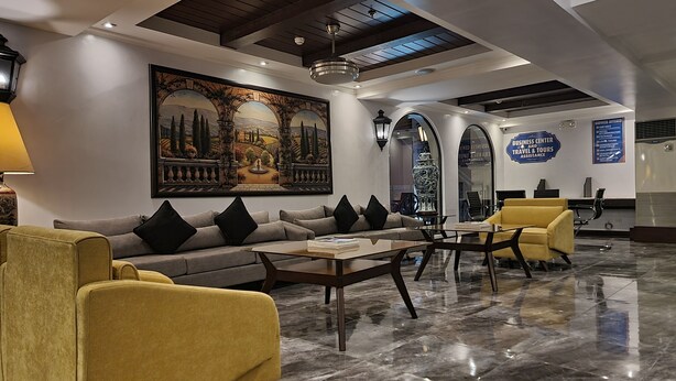 A lobby with a sofa, armchairs, a coffee table, a large painting, and a sign for business center and travel assistance. - Royal Bellagio Hotel (Makati)