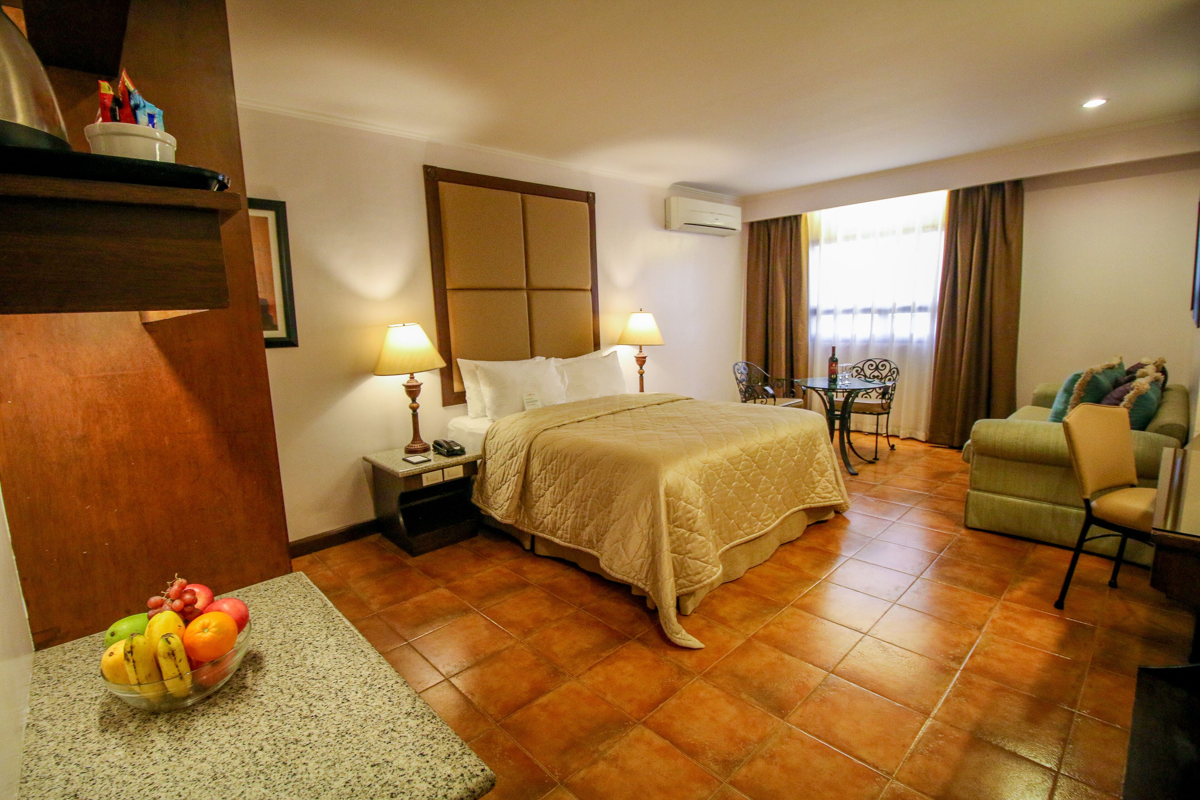 Quarto de hotel com cama, luminárias de cabeceira, uma mesinha com cadeira, um sofá e uma cesta de frutas no chão.