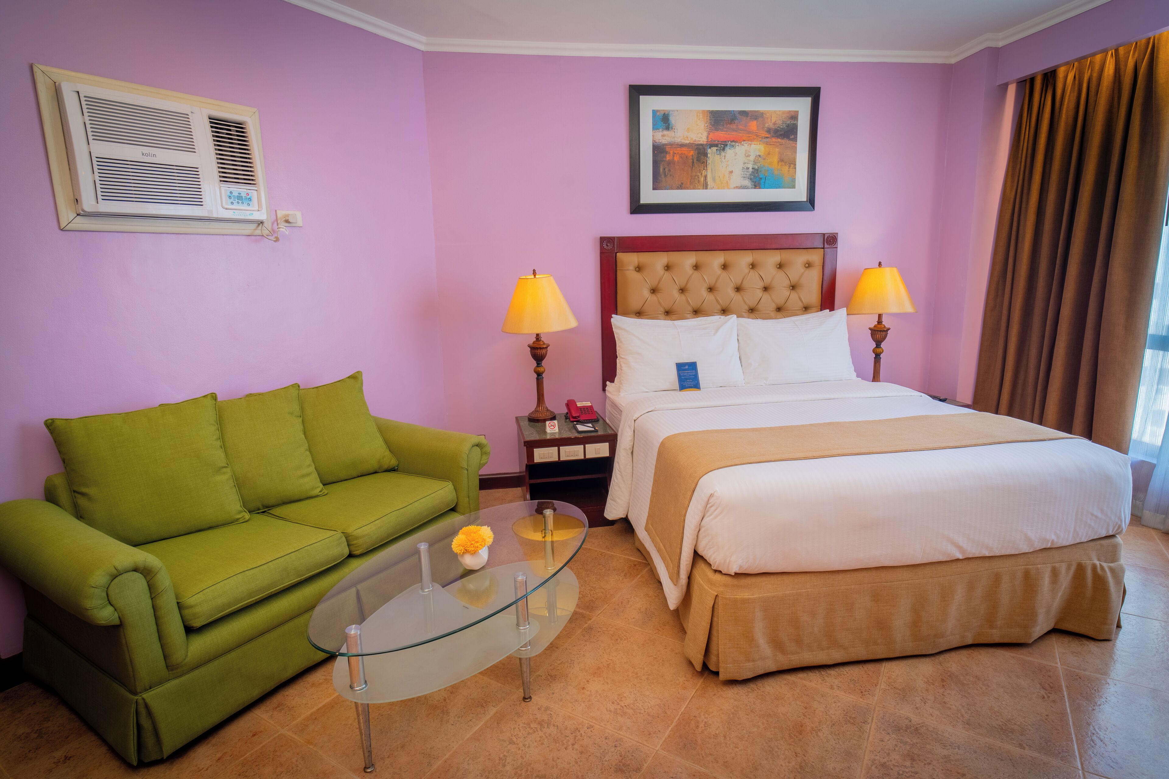 Une chambre d’hôtel avec un canapé vert, une table basse en verre ornée d’une fleur, un lit avec une literie blanche, deux lampes de chevet, une œuvre d’art encadrée au mur et un appareil de climatisation.