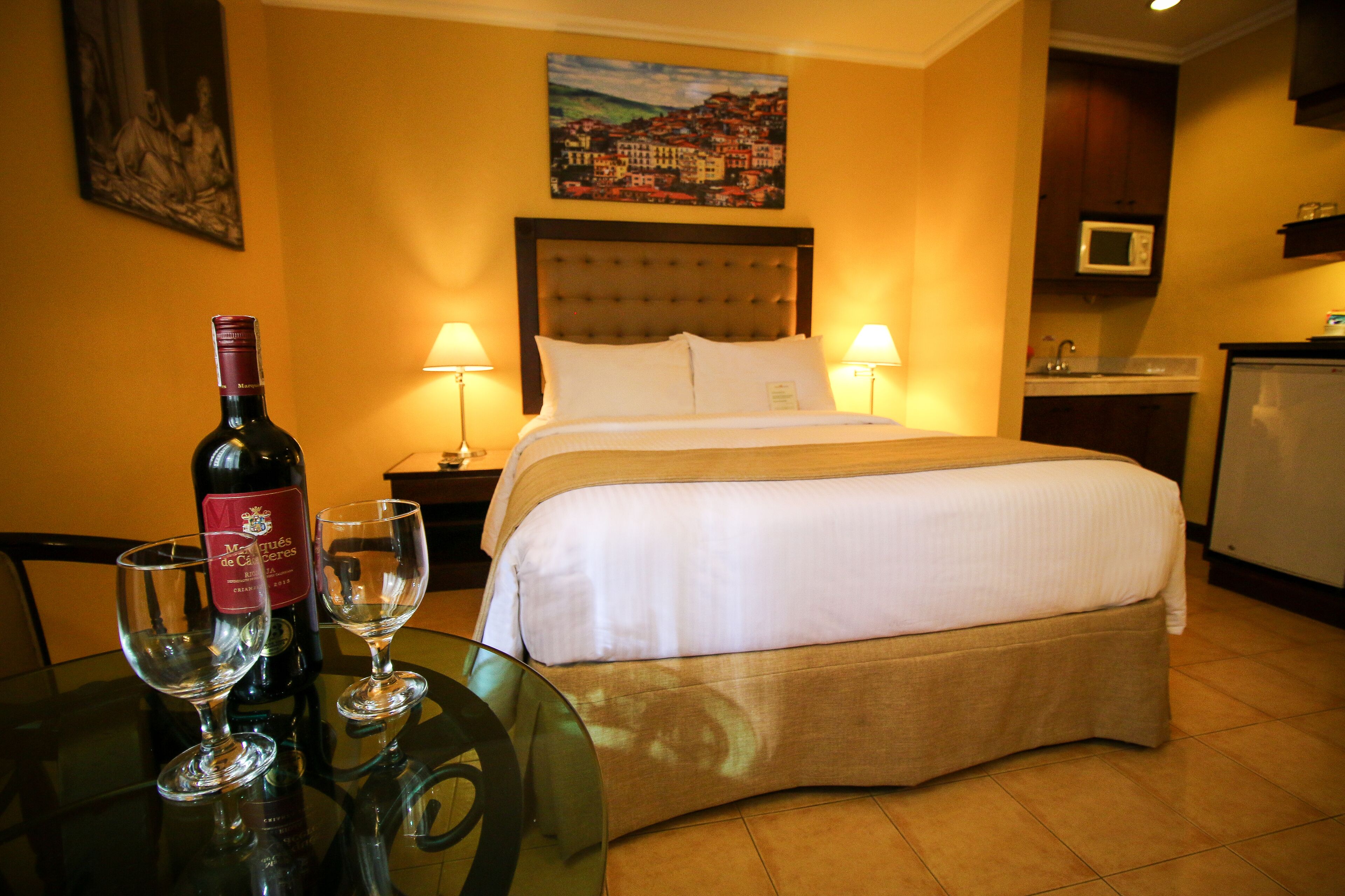 A hotel room with a bed, a glass table with wine glasses and a bottle, a painting on the wall, and a kitchen area.
