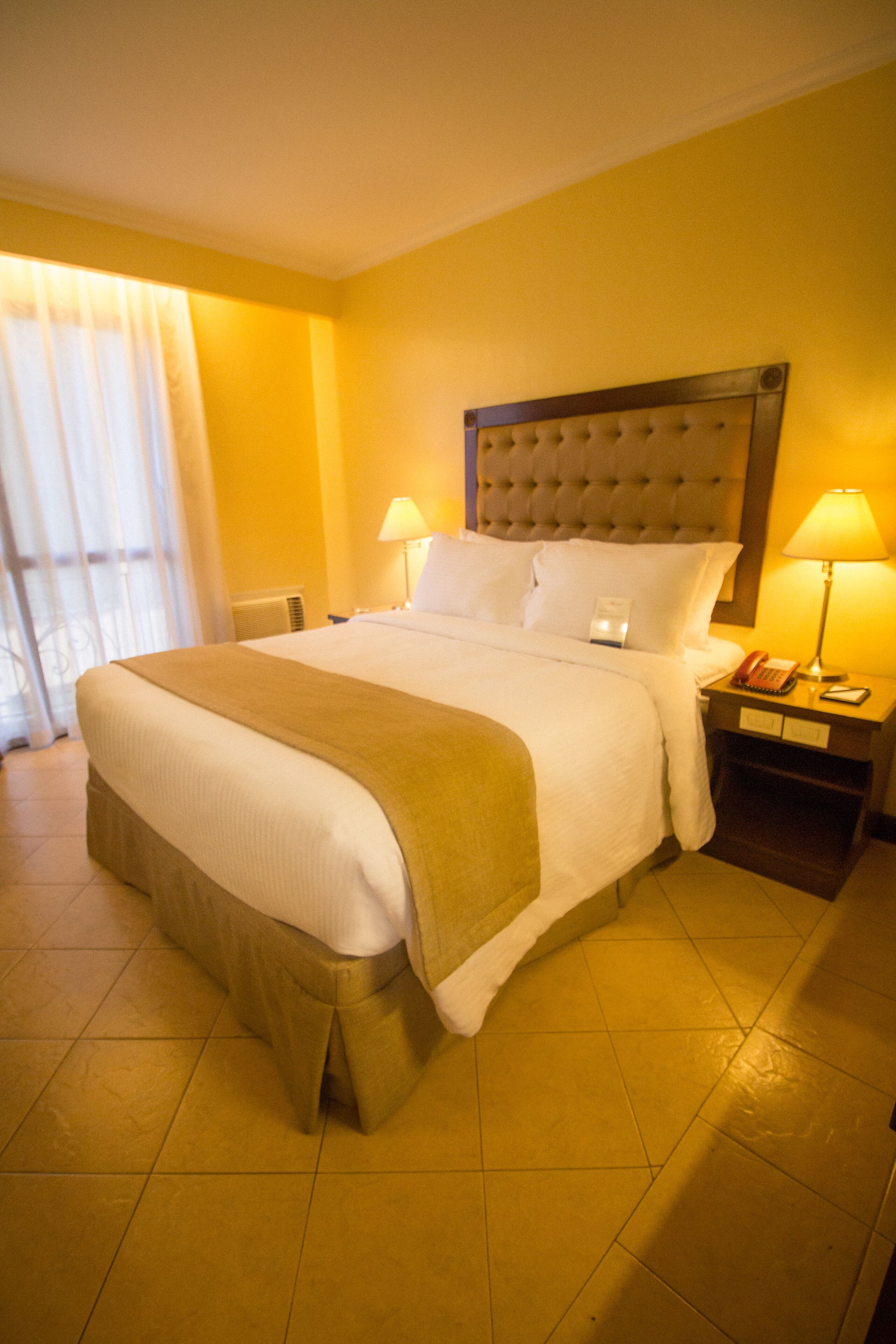 Habitación de hotel con una cama grande, dos mesitas de noche, cabecero y una ventana con cortinas.