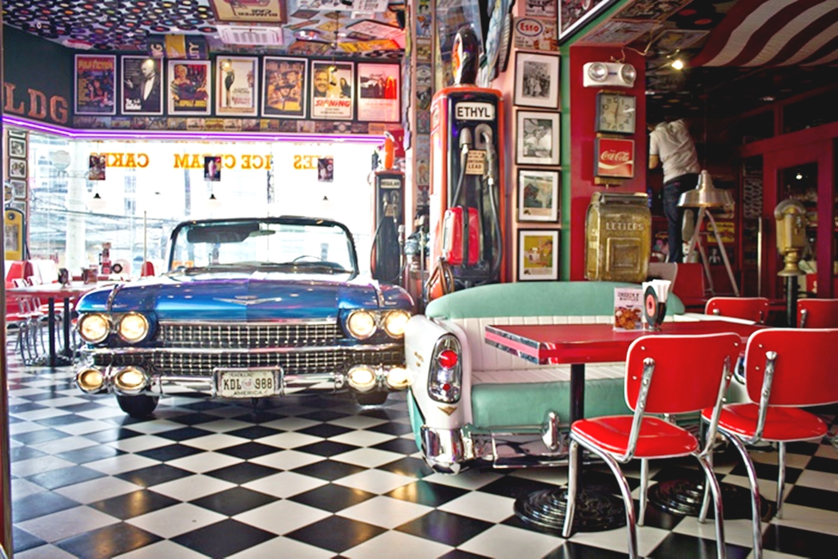 Un interior de restaurante con coches antiguos, piso a cuadros, letreros de neón y decoración retro.