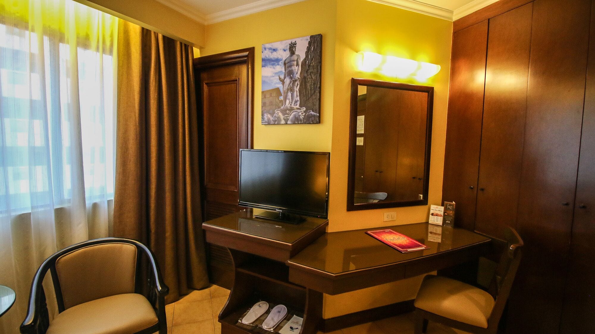 A hotel room with a desk, television, chairs, a mirror, and a window with curtains.