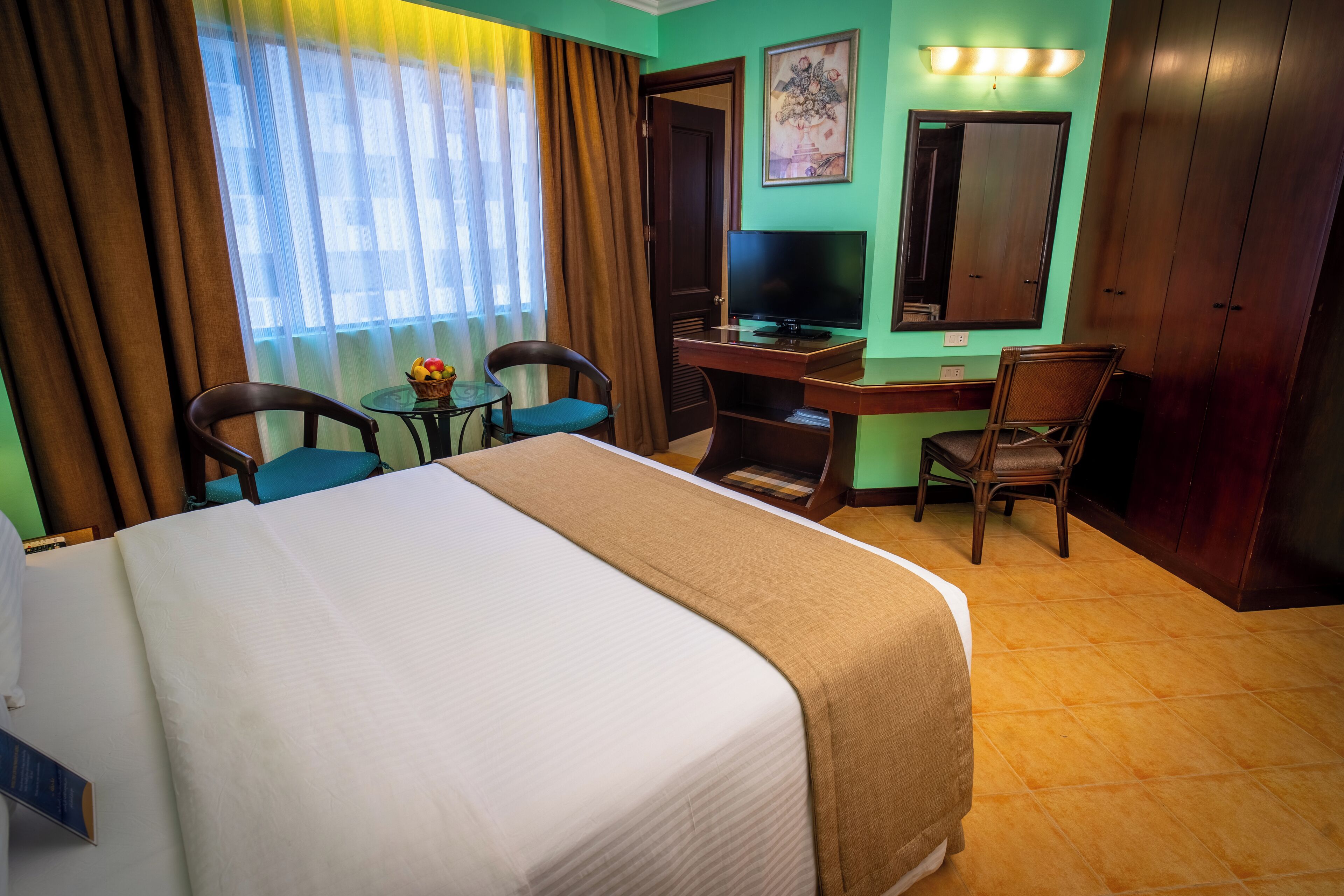 A hotel room with a large bed, a desk with a computer, two chairs, a small table with fruit, and a TV.