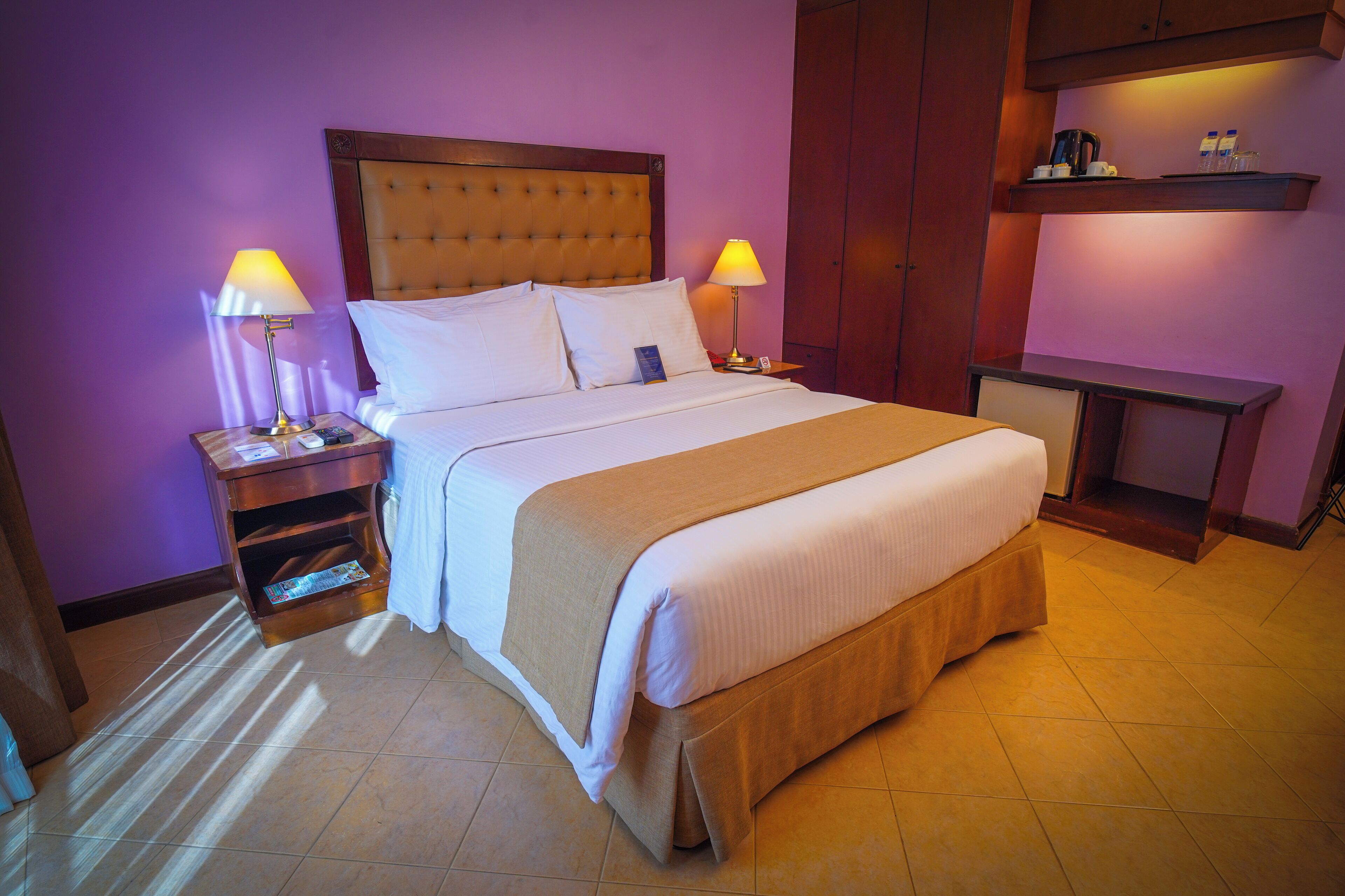 Una cama bien hecha con cabecera, dos mesitas de noche con lámparas, un tocador de madera y una pequeña estantería con objetos.