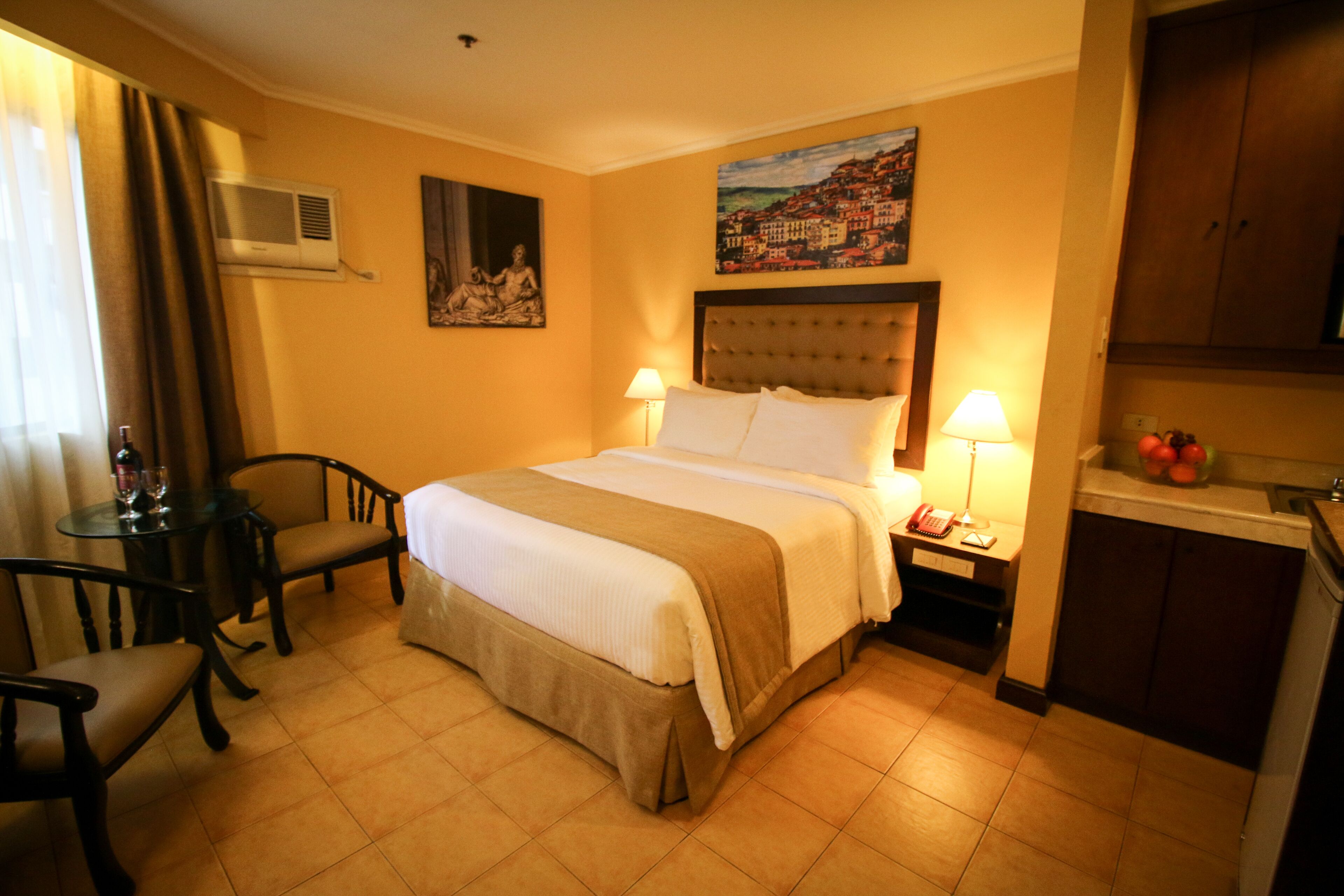A hotel room with a bed, two bedside tables with lamps, a small table with chairs, a painting on the wall, and a kitchen area with a sink and fruit bowl.