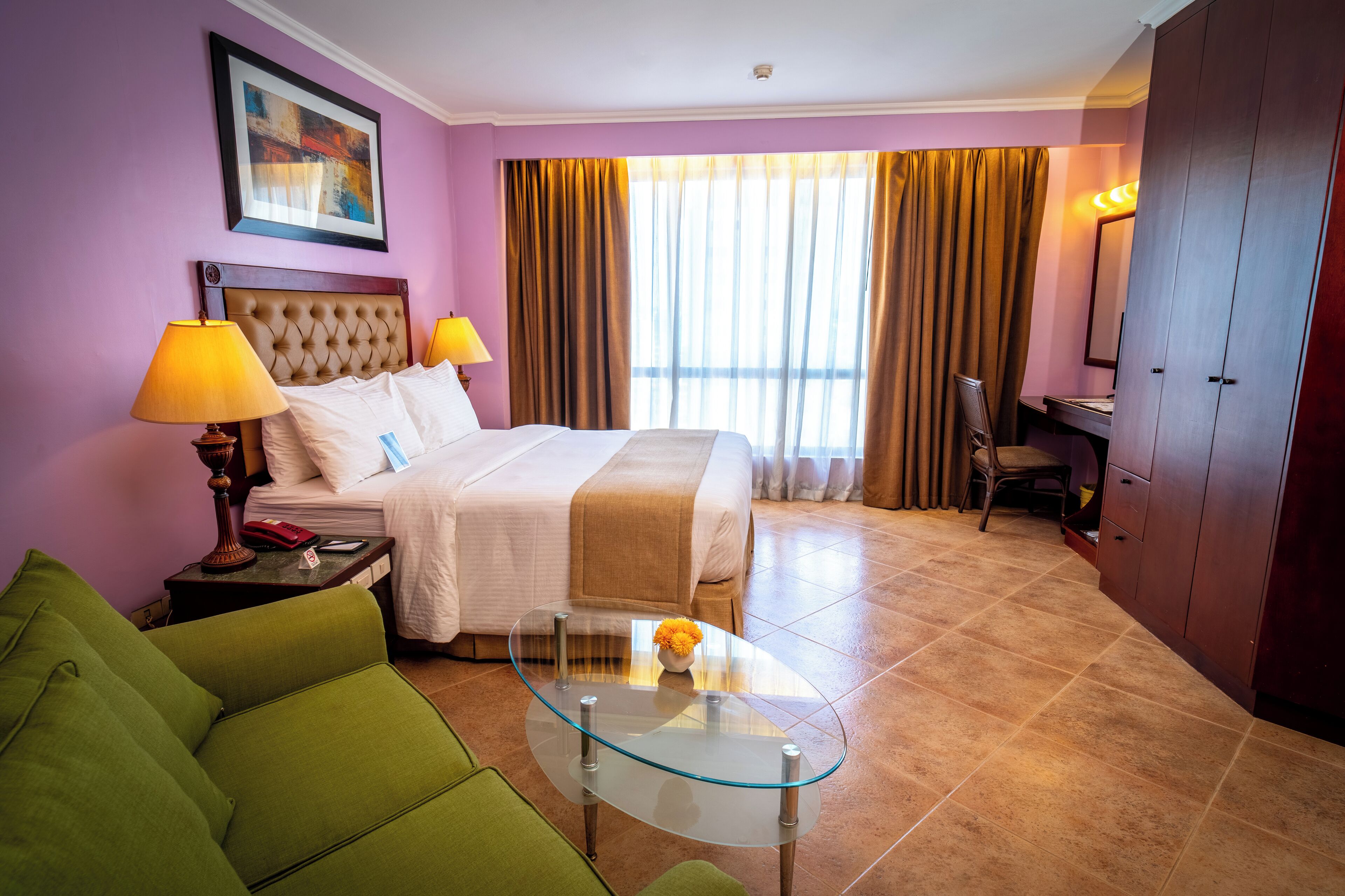 A hotel room with a bed, a green sofa, a glass coffee table, a desk with a chair, and a wardrobe.