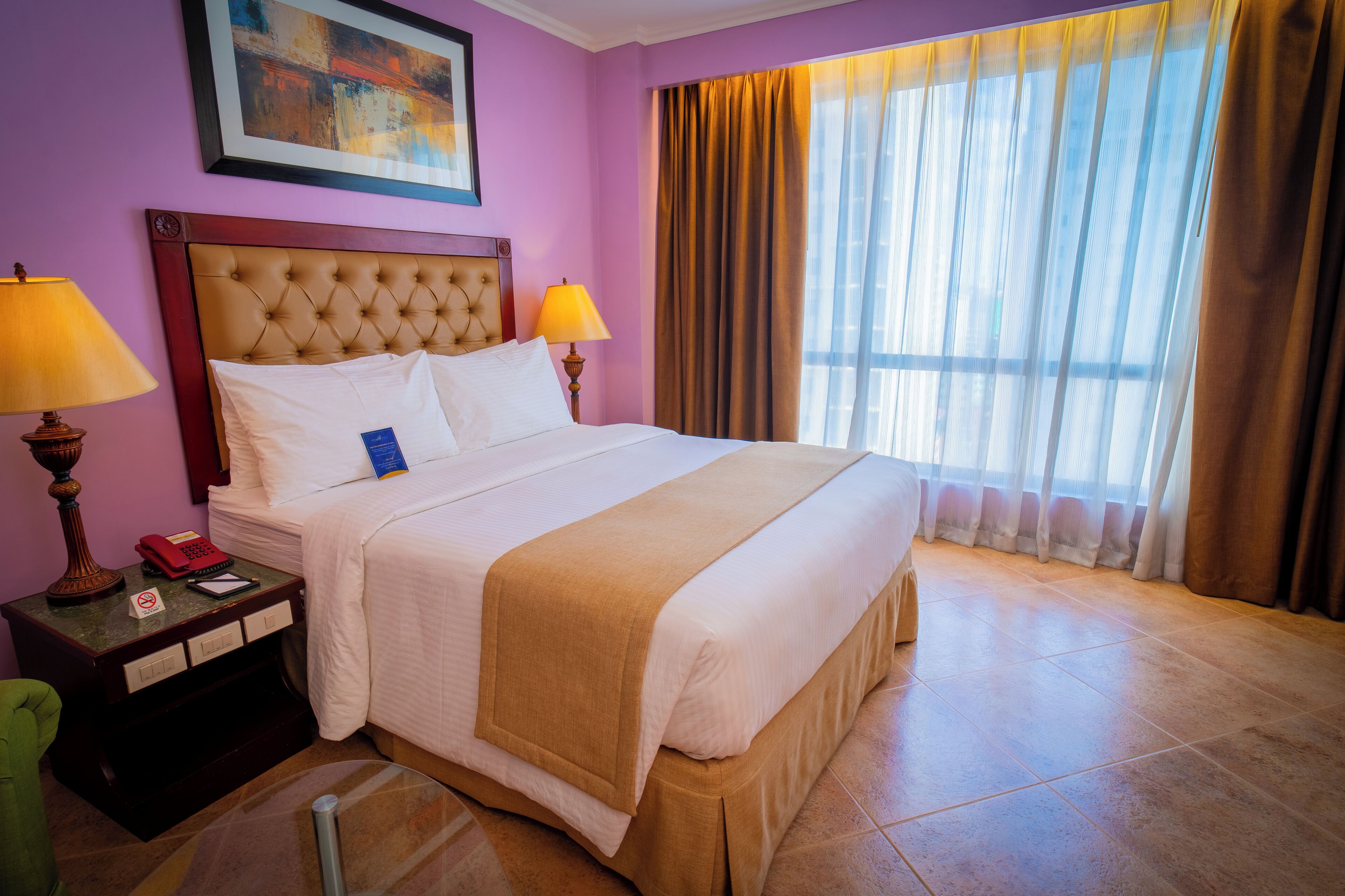 A hotel room with a large bed, two bedside lamps, a nightstand with a telephone and a notepad, a green chair, and a window with curtains.