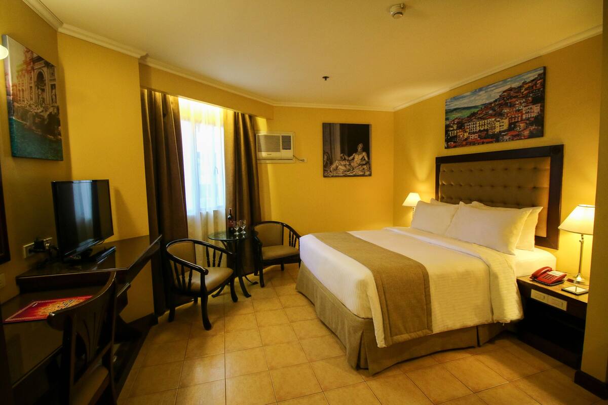 A hotel room with a bed, a desk with a chair, a television, and a painting on the wall.