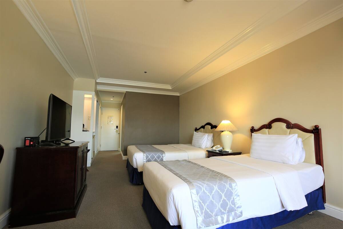 A hotel room with two beds, a television on a dresser, and a desk with a lamp.