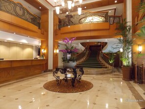 A grand lobby with a reception desk, a staircase, and decorative plants.