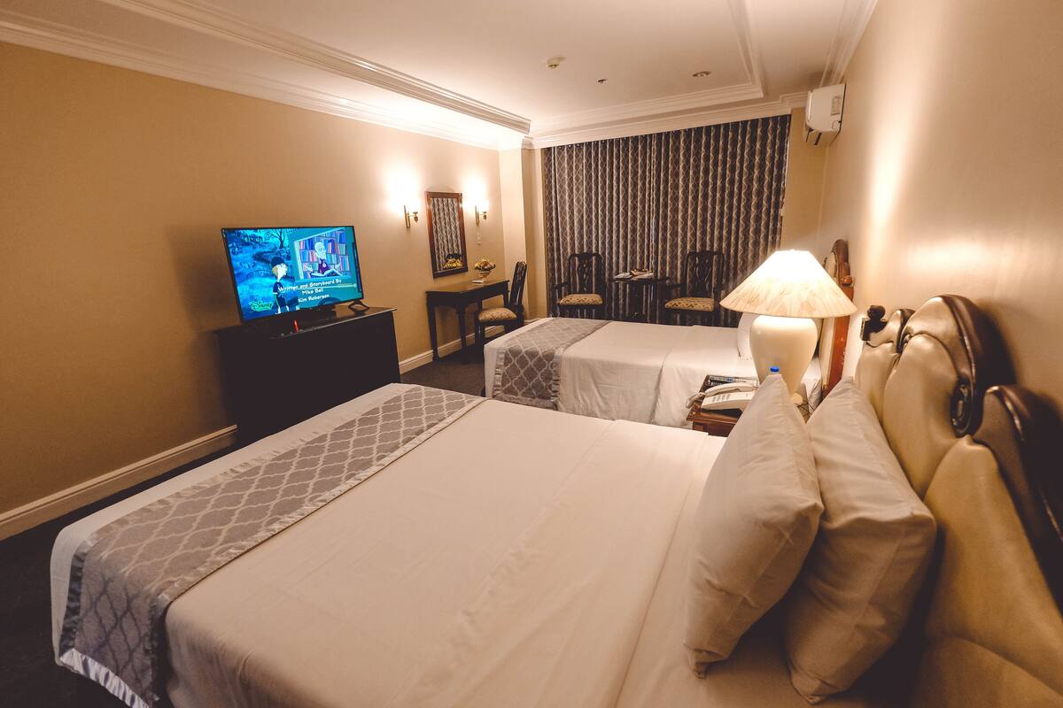 A hotel room with two beds, a TV, a desk with a chair, and a lamp.