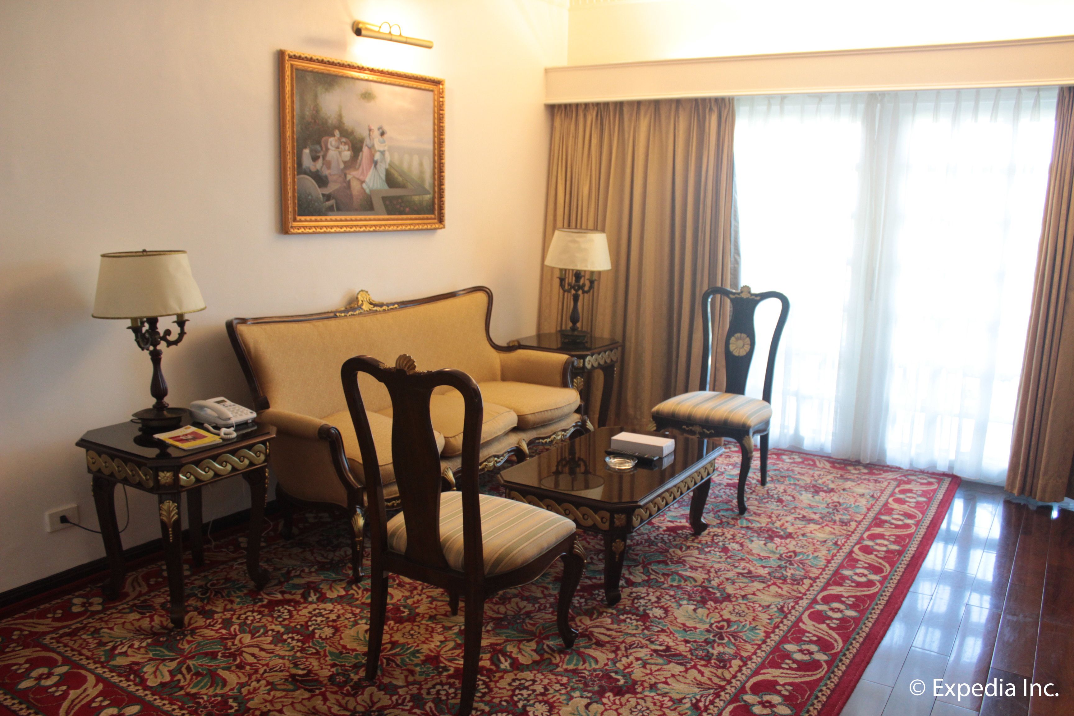 Habitación de hotel con sofá, dos sillones, una mesita, lámpara, un cuadro y un ventanal con cortinas.