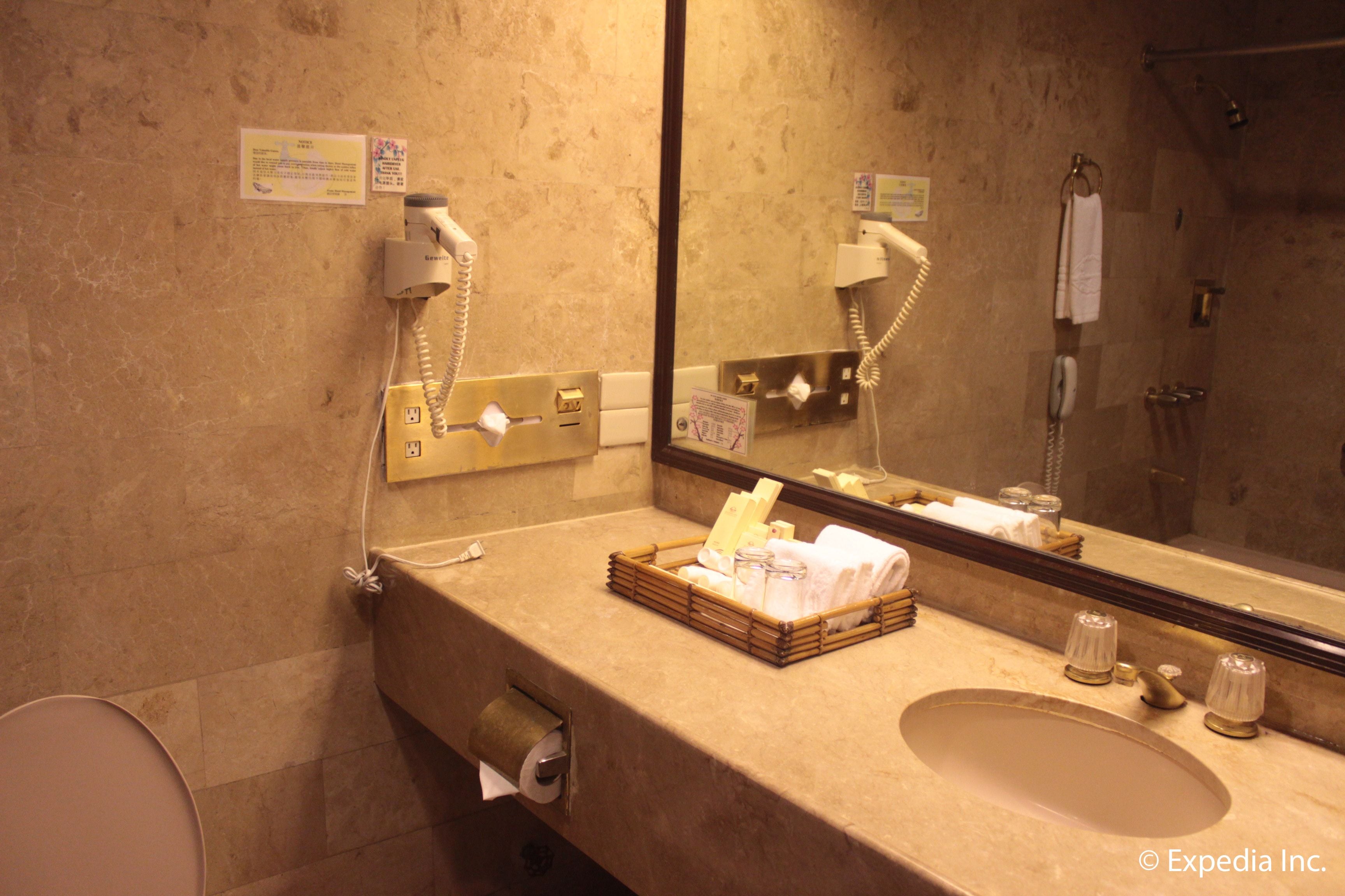 Une salle de bain équipée d’un lavabo, d’un miroir et de deux téléphones. On y trouve un panier avec des serviettes et un rouleau de papier toilette.