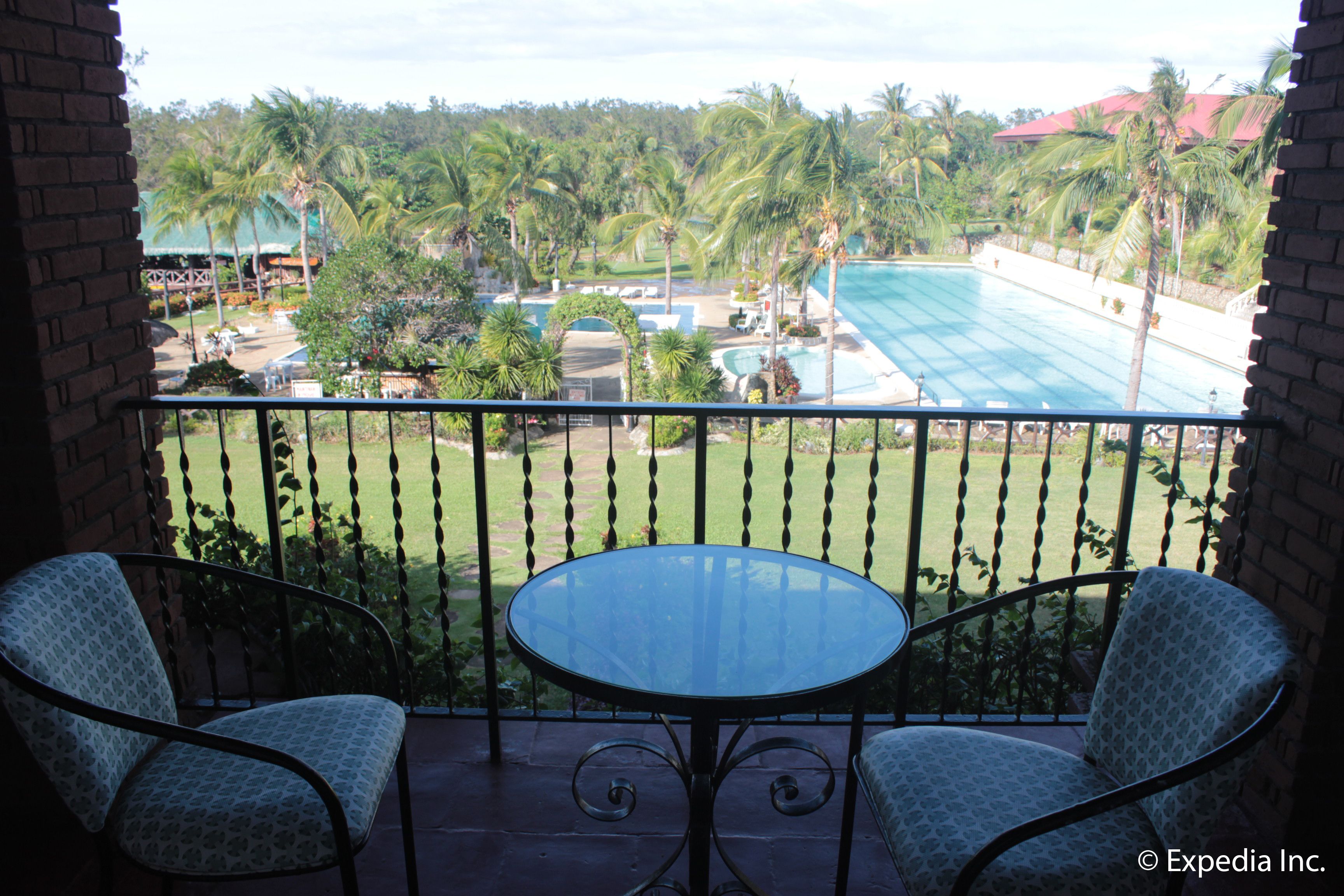 A balcony with two patterned chairs and a glass table, overlooking a swimming pool and lush greenery.