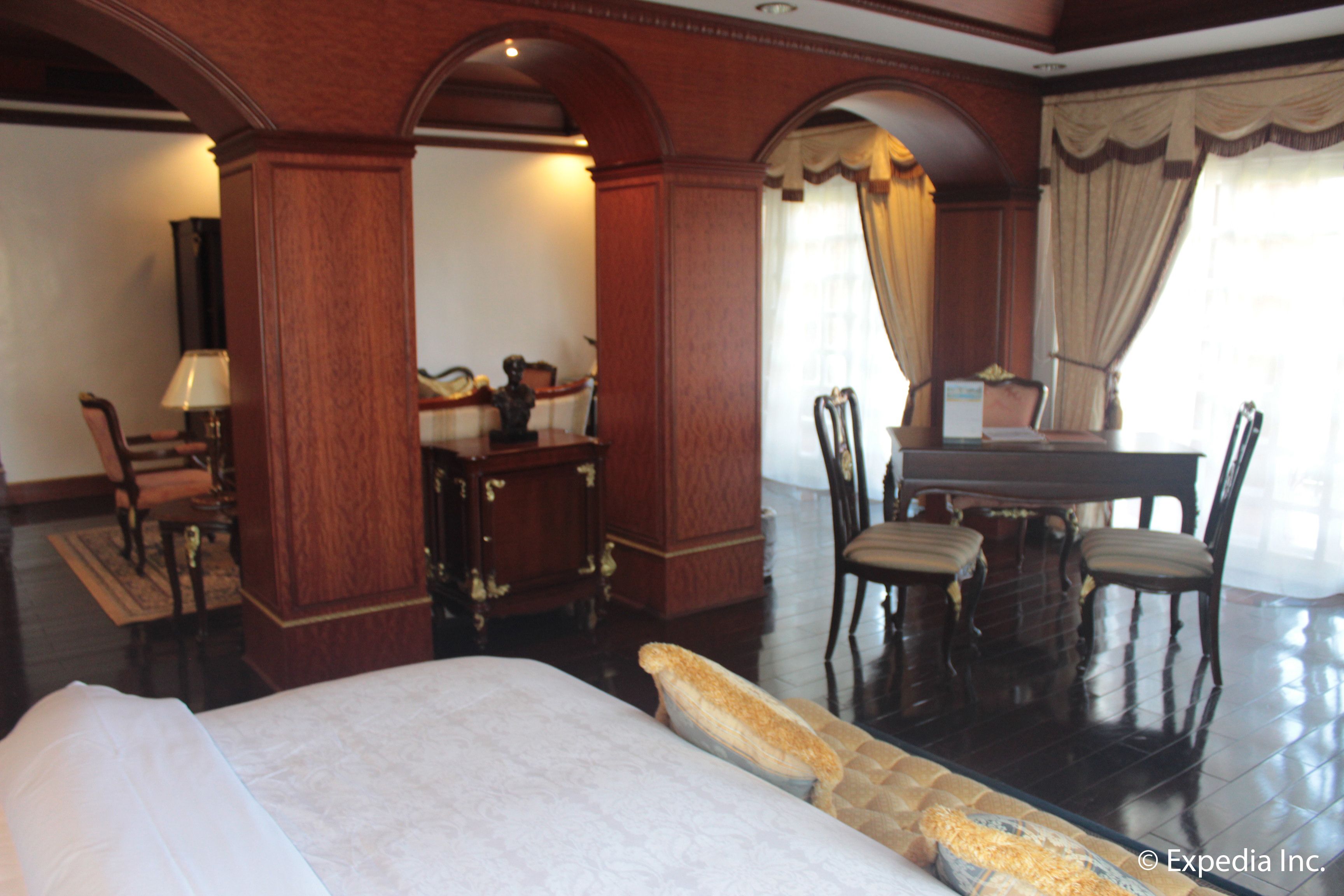A hotel room with a large bed, a dining table with chairs, a desk with a chair, and a piano.