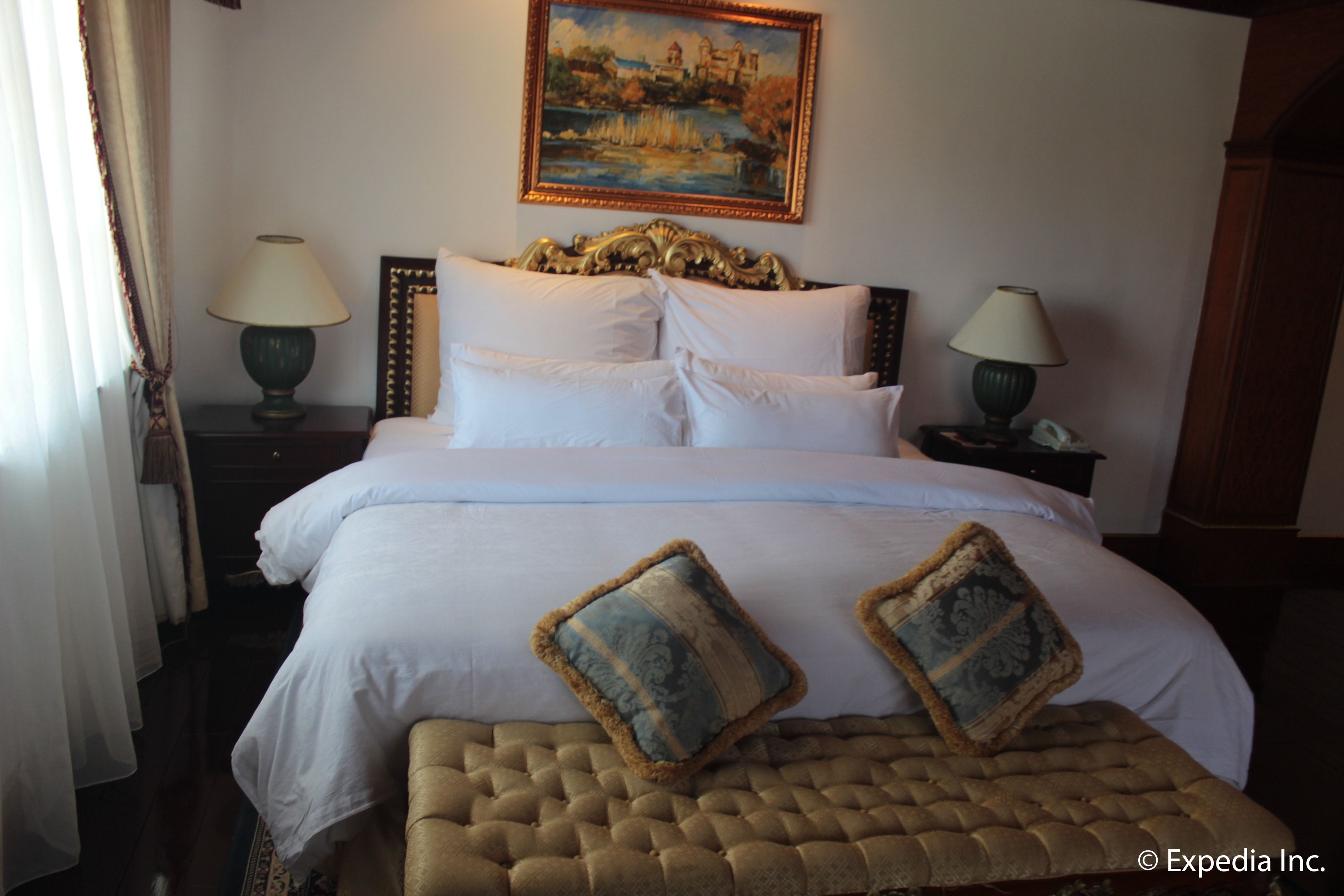 A hotel room with a large bed, two bedside lamps, a painting on the wall, and a wooden bench.
