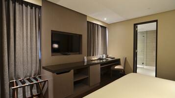 A hotel room with a bed, a desk with a flat-screen TV, a chair, a shower area, and a towel rack.