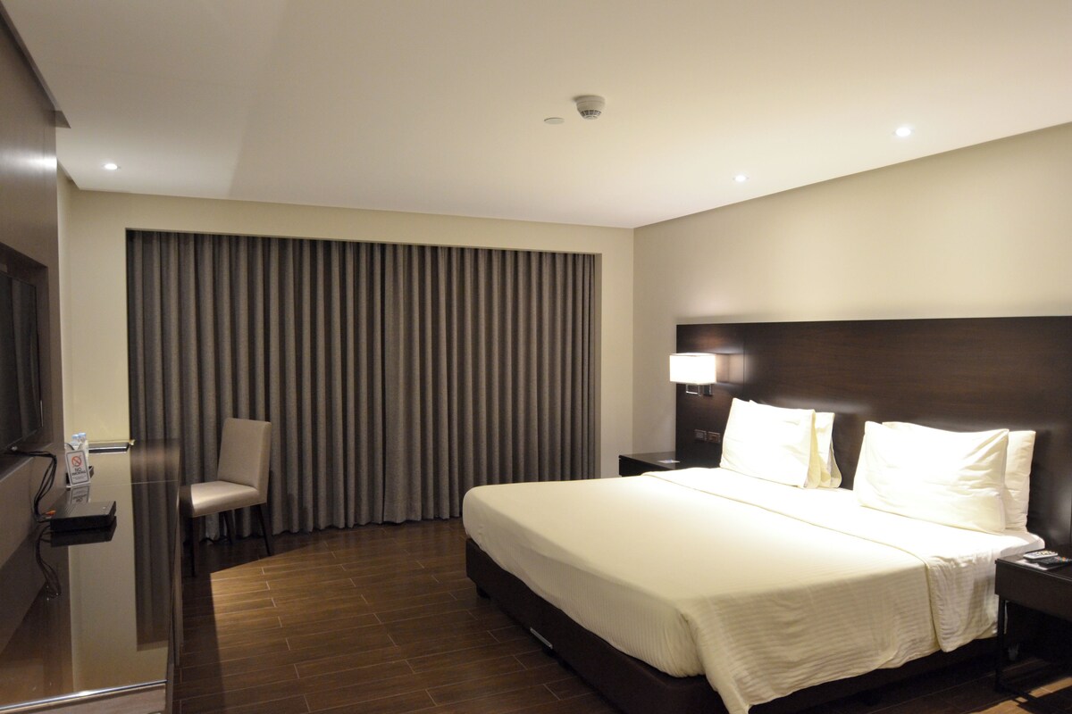 A hotel room with a large bed, a bedside table, a chair, a desk with a computer, and a window with curtains.