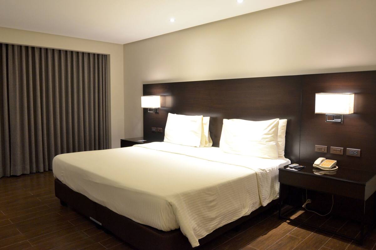A hotel room with a large bed, two bedside lamps, a nightstand with a phone, and a window with curtains.