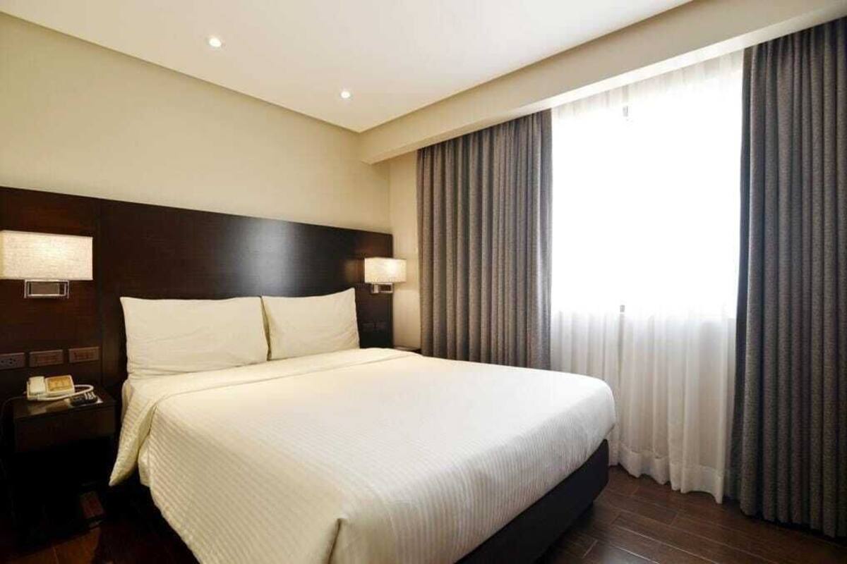 A hotel room with a large bed, two bedside lamps, a nightstand with a phone, and a window with curtains.
