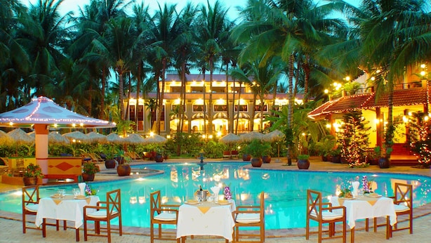 A poolside dining area with tables and chairs, surrounded by palm trees and a building with illuminated windows. - Vinh Suong Seaside (Mui Ne)