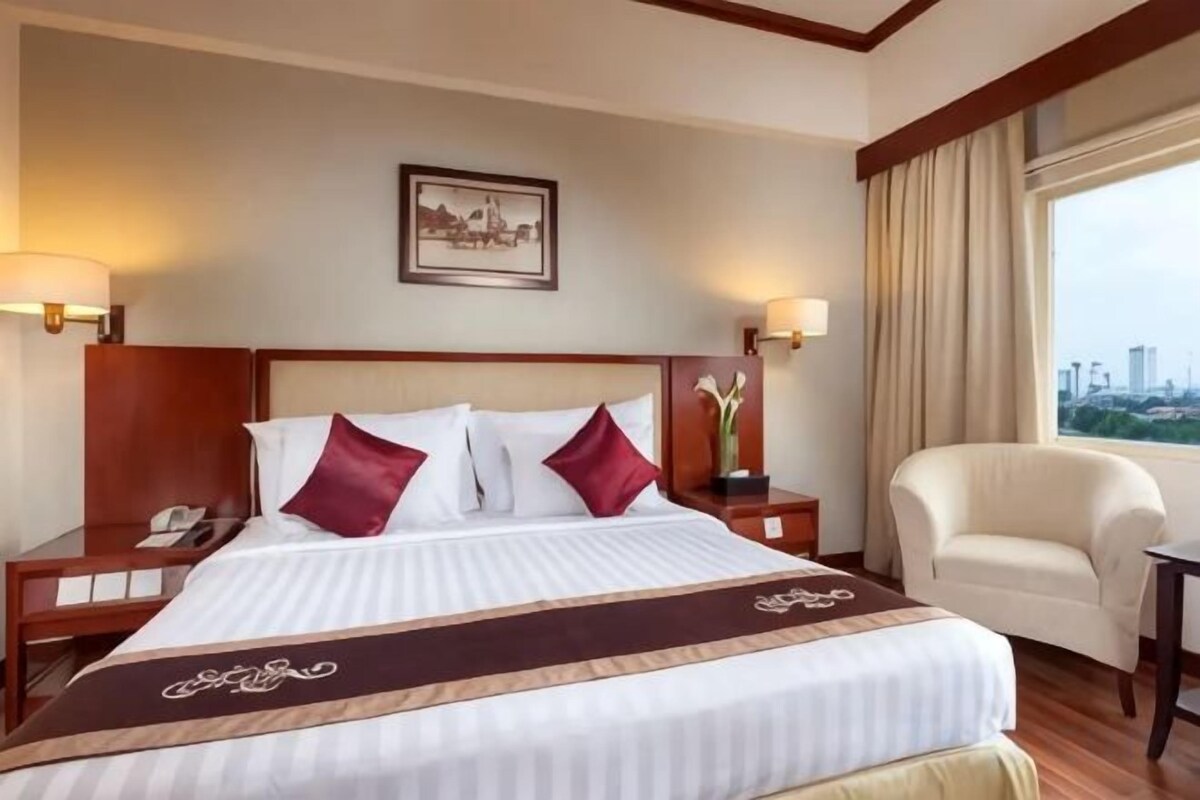 A hotel room with a large bed, two bedside tables, a chair, a desk, and a window with a city view.