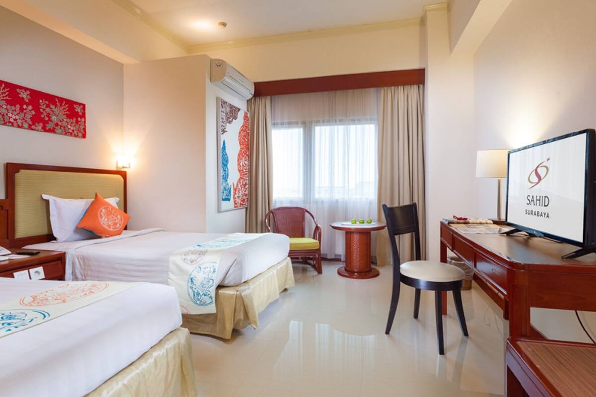 A hotel room with two beds, a desk, a chair, a TV, and a window with curtains.