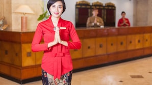 Uma área de recepção de hotel com recepcionista de uniforme vermelho, balcão de madeira e uma planta ao fundo.