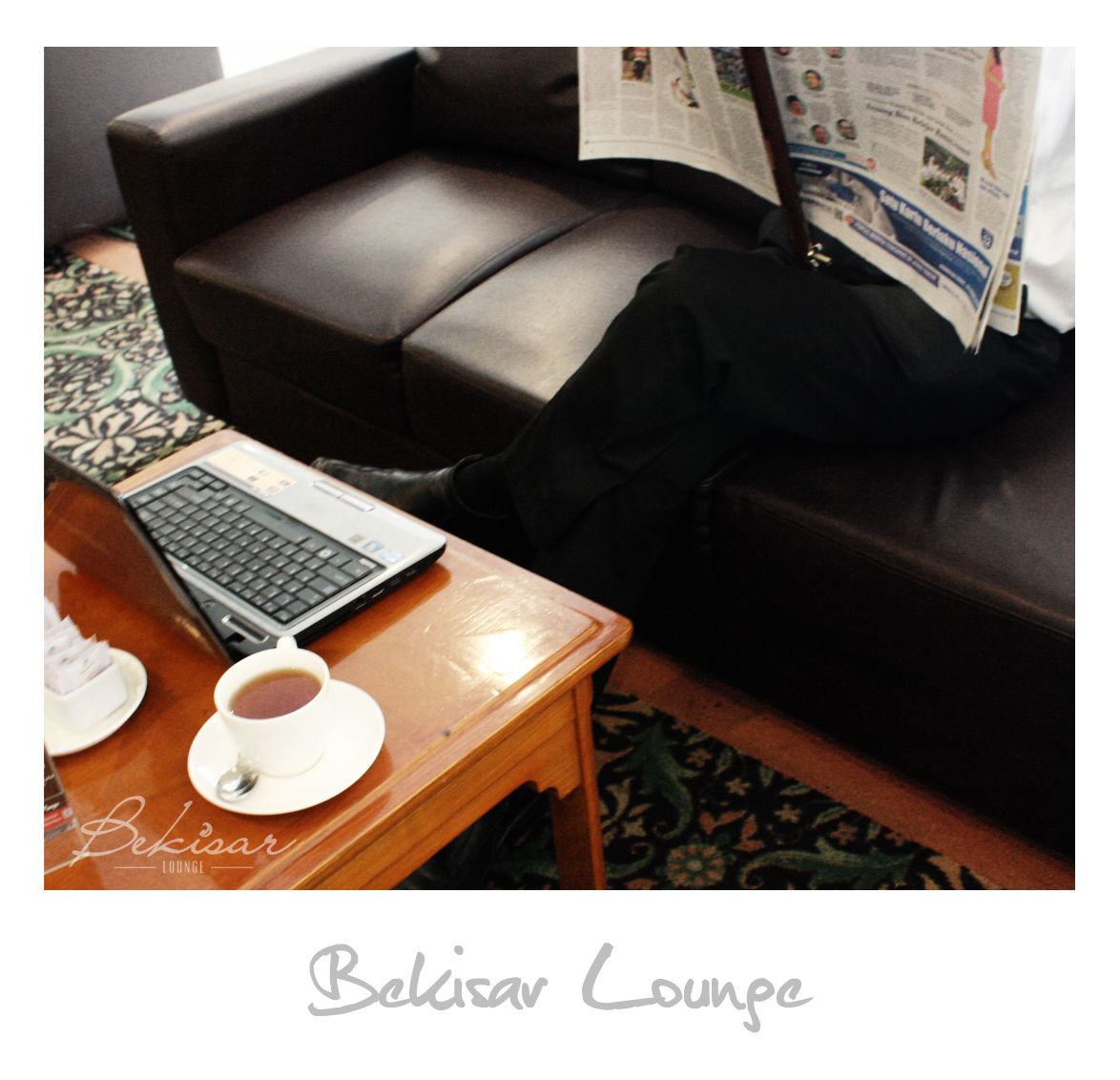 Een Belisar Lounge met een leren bank, een houten tafel met een laptop en een kop thee, en een kamerbreed tapijt met dessin.