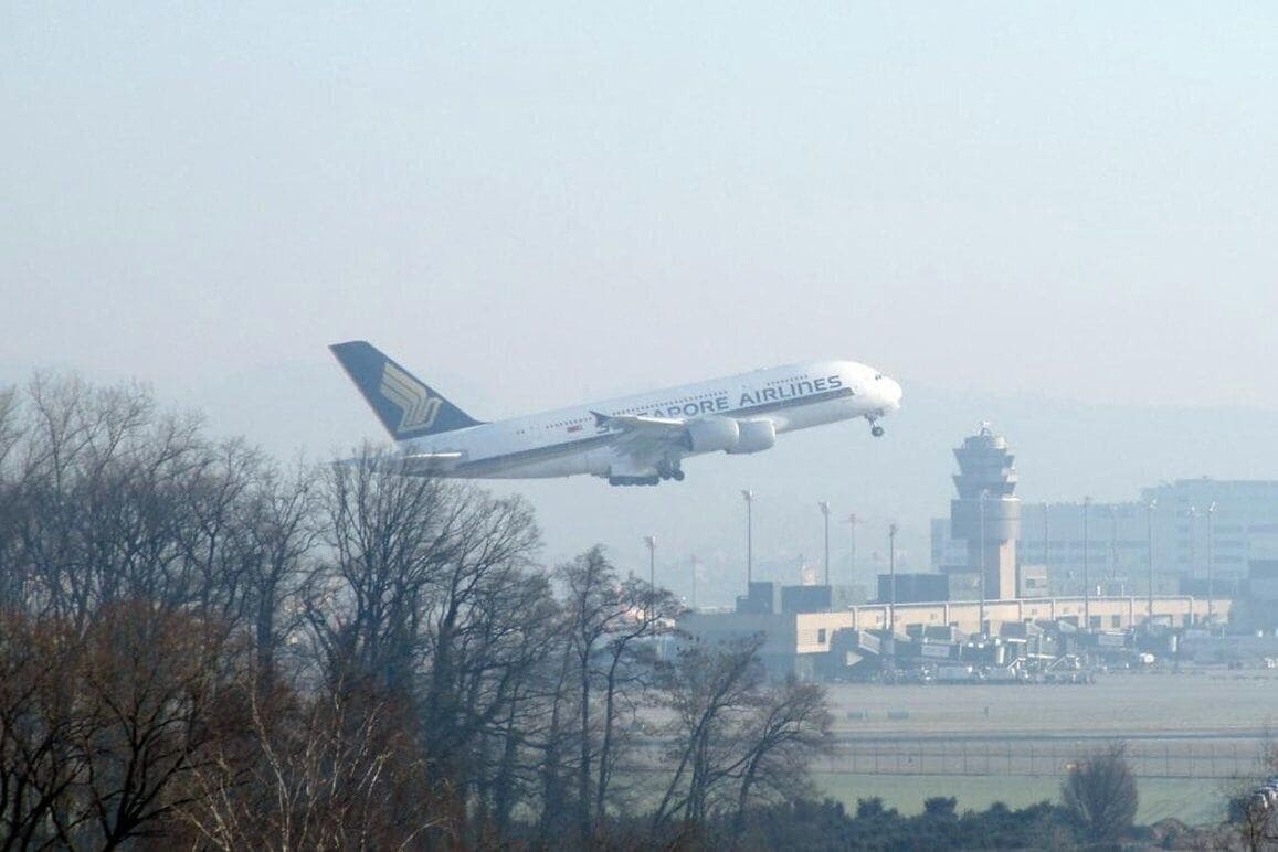 An airplane with 'SINGAPORE AIRLINES' on its side flying over a landscape with trees and a building in the background.