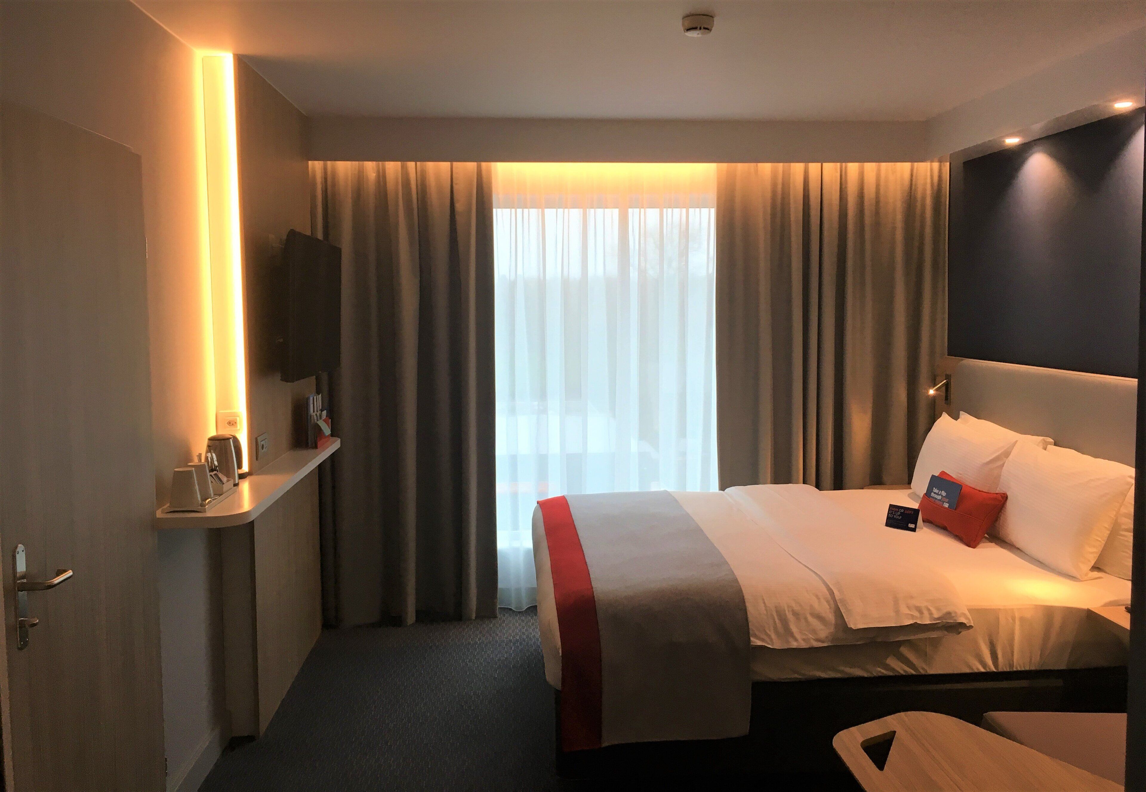 Une chambre d’hôtel avec un lit, une table de chevet, une télévision et une fenêtre avec des rideaux.